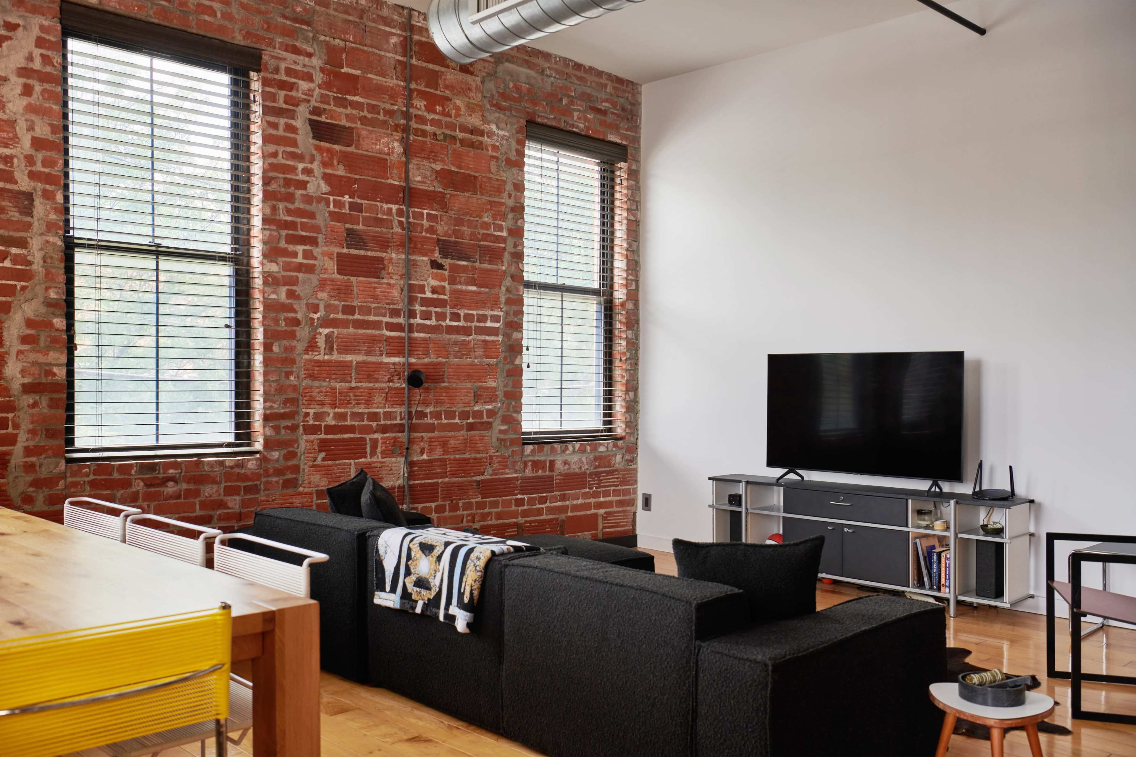 A living room features exposed brick walls, a black sectional sofa, a television on a media console, and large windows with blinds.