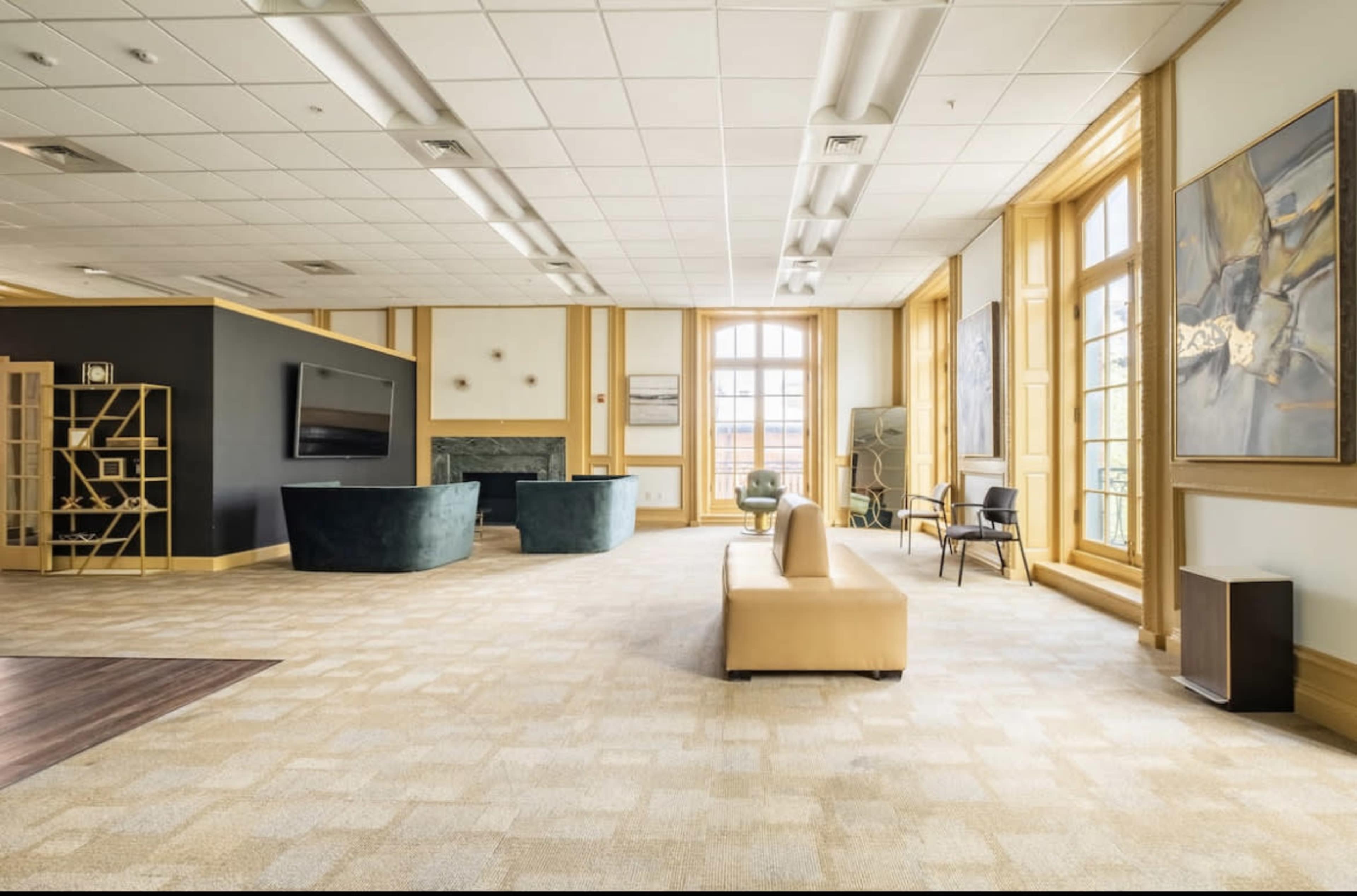 A spacious lounge area featuring a beige sofa, a dark green seating arrangement, a large television on the wall, and large windows allowing natural light to fill the room.