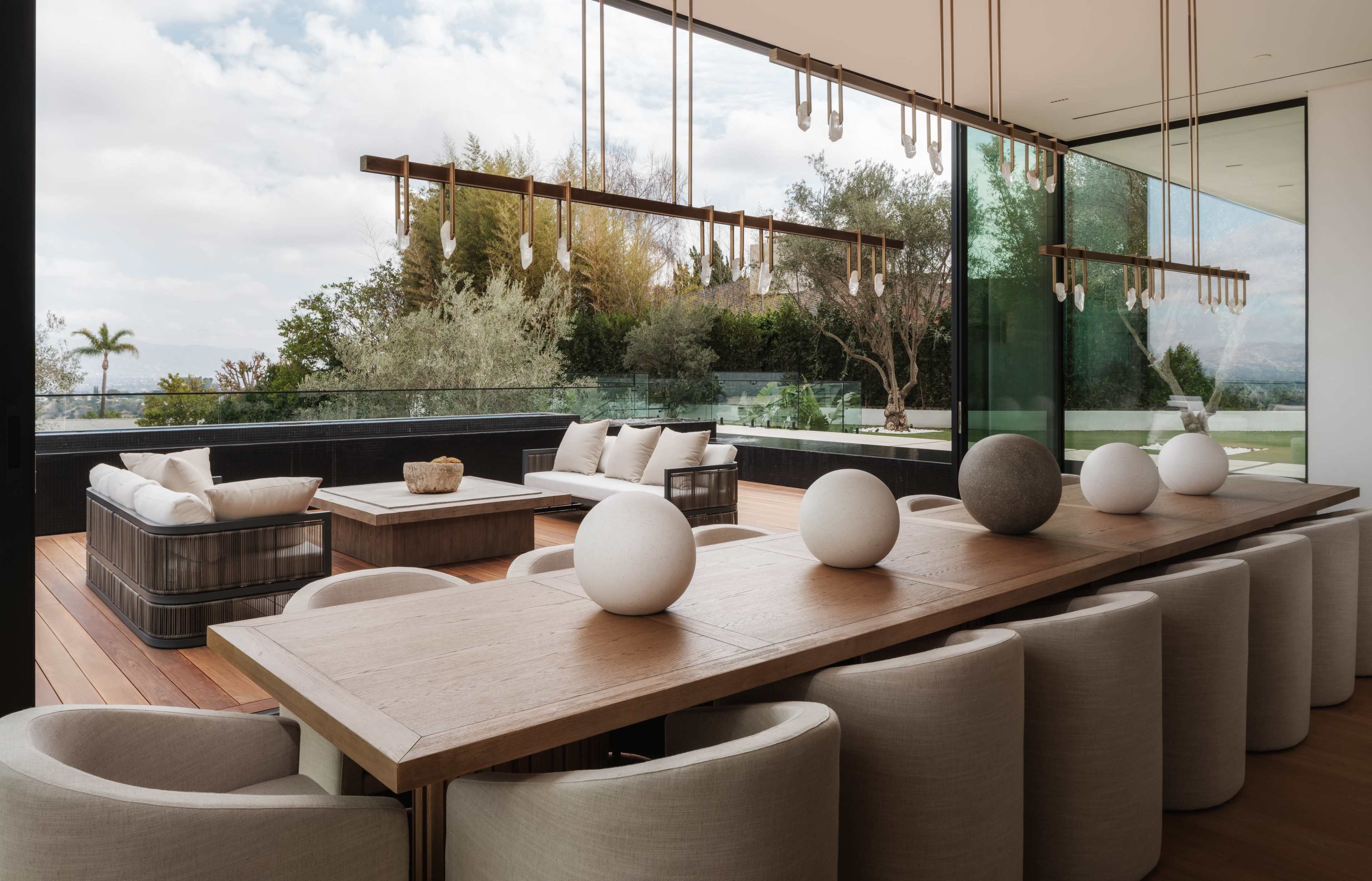 A modern dining area features a long wooden table with circular decorative objects, surrounded by upholstered chairs and large windows overlooking an outdoor space with greenery.