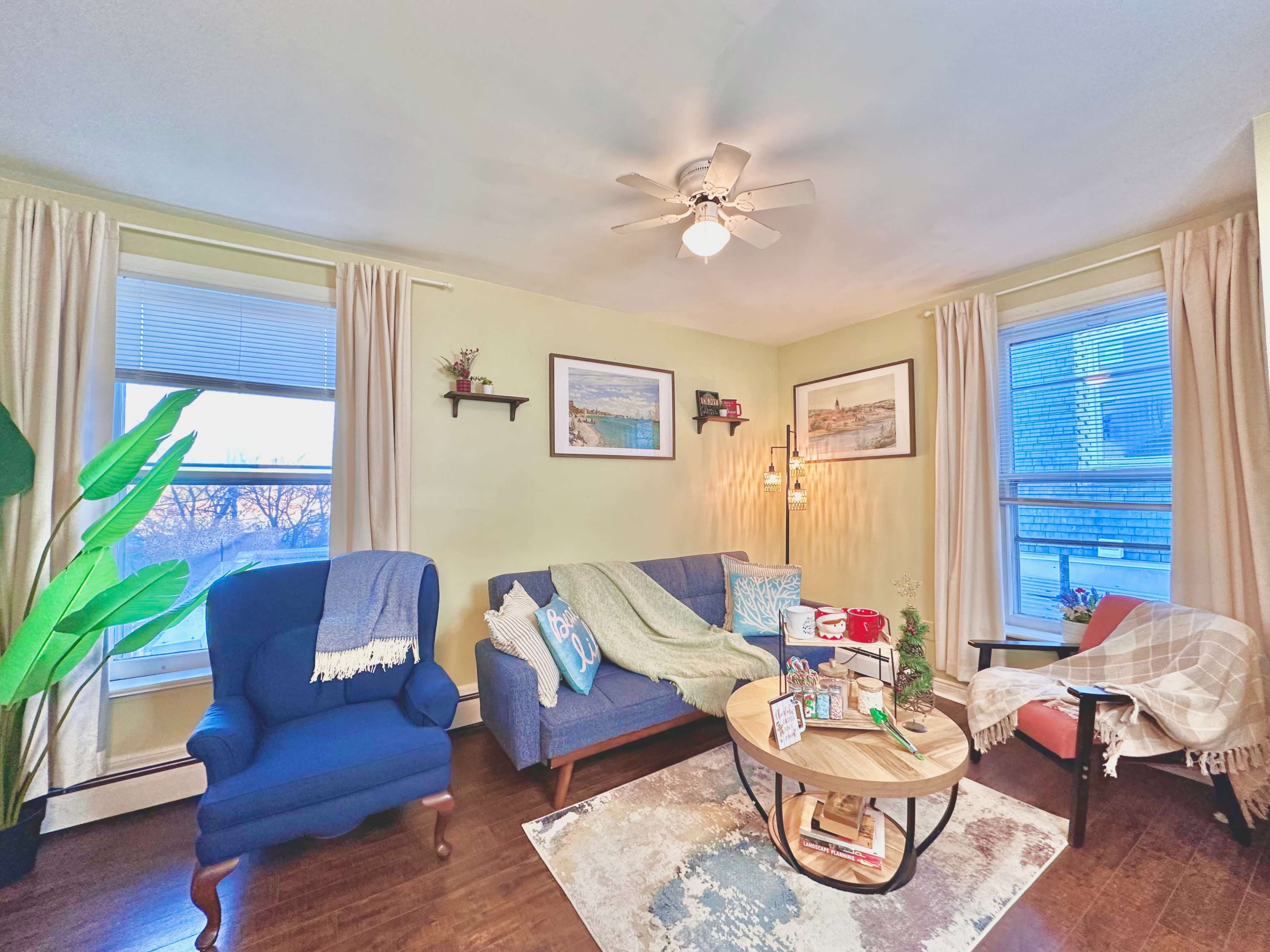 The image shows a cozy living room with a blue sofa, a round coffee table, and large windows dressed in light curtains.