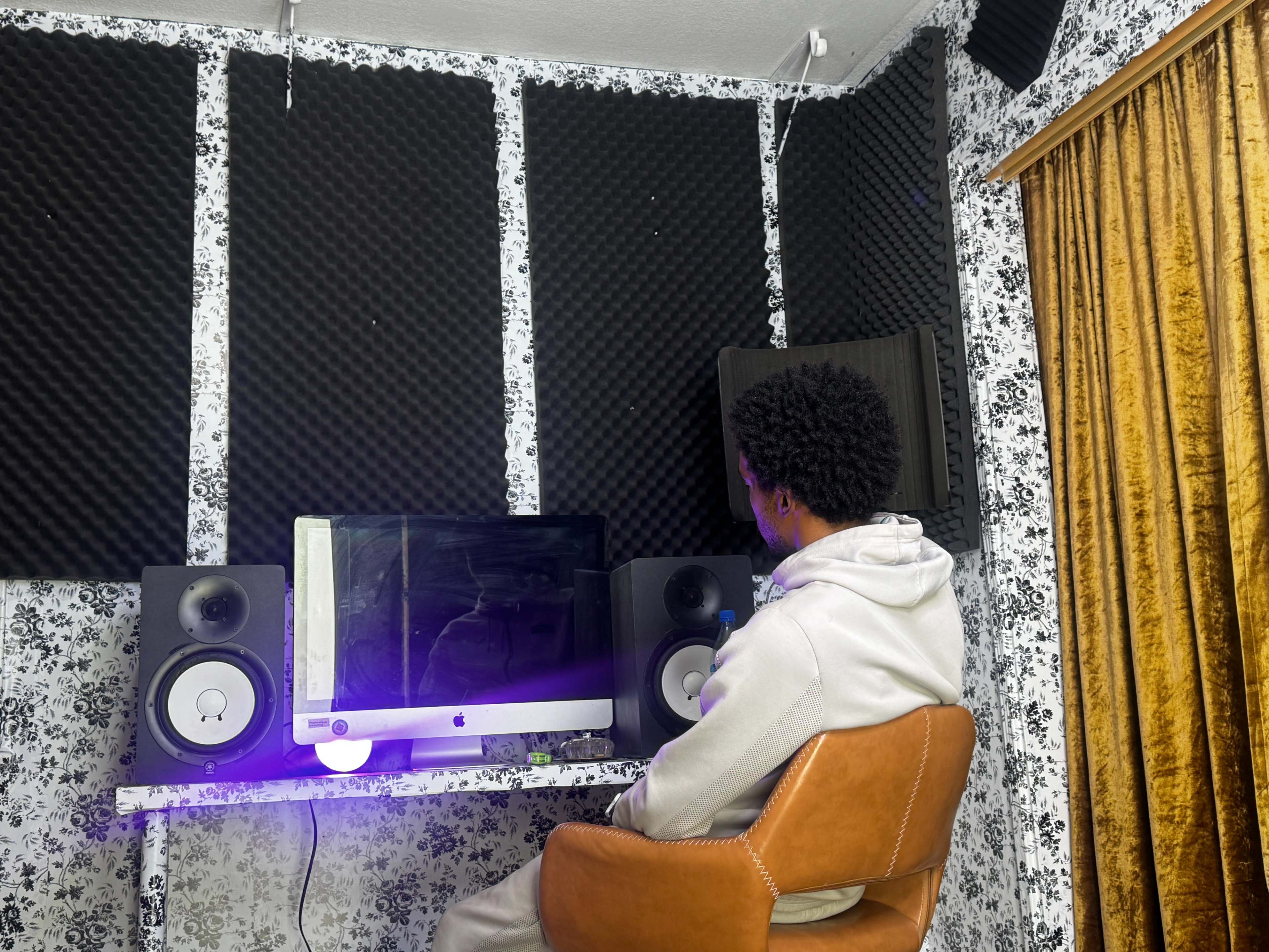 A person sits in an orange chair in front of a computer, surrounded by soundproofing panels in a room with patterned wallpaper and curtains.