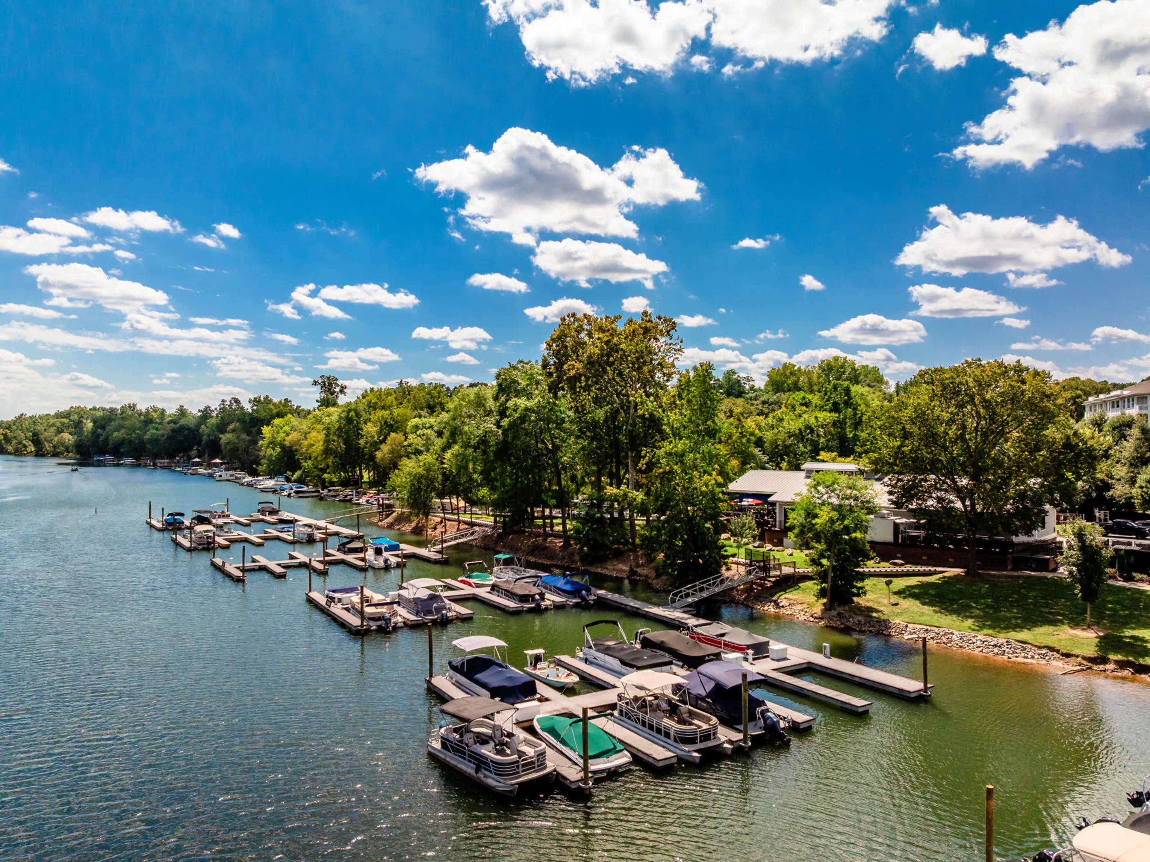 The image shows a marina on a tranquil lake, featuring several boats docked at wooden piers under a clear blue sky with scattered clouds.