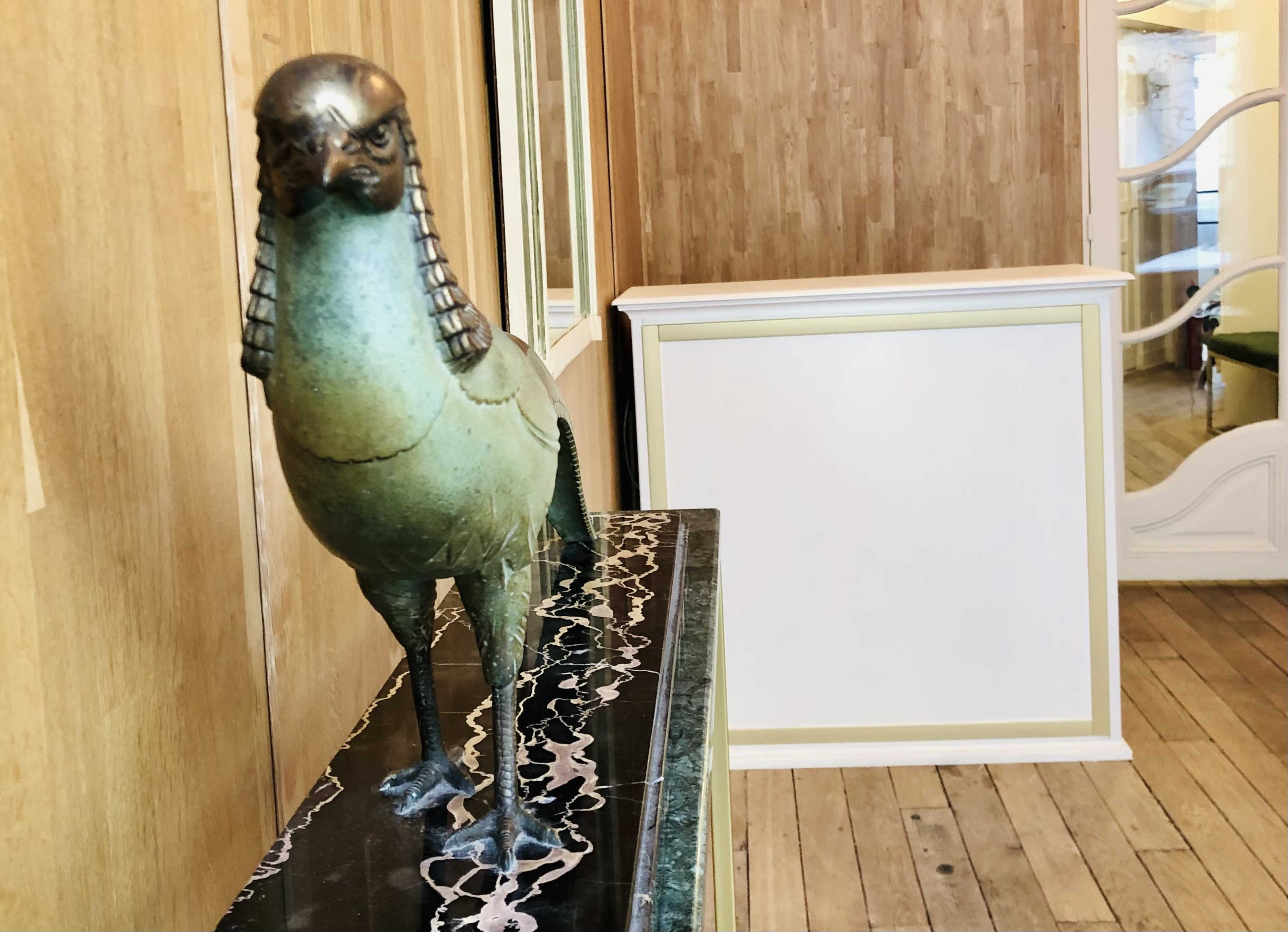 A bronze bird sculpture stands on a dark surface in a wooden-paneled room with a blank white wall in the background.