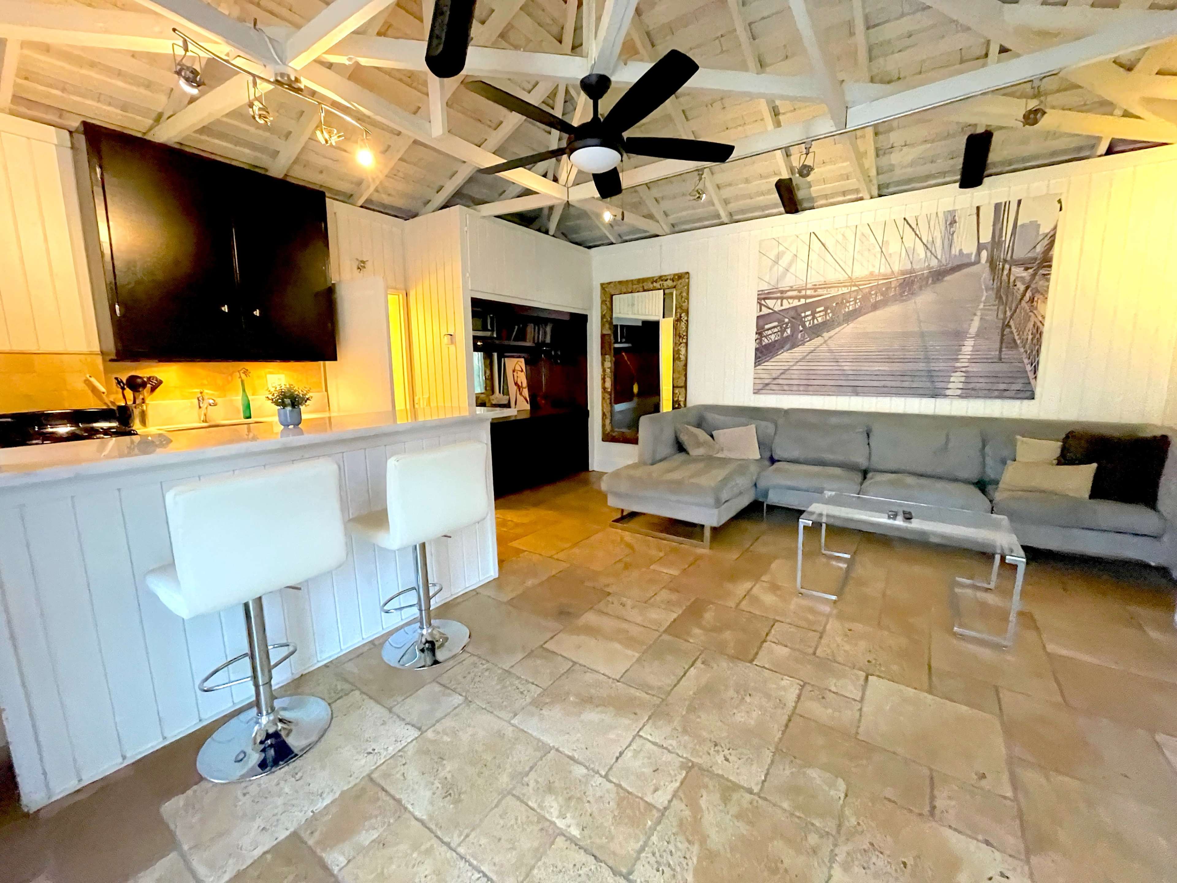 The image shows a spacious interior with a kitchen area featuring bar stools, a living area with a gray sofa, and a large wall art piece depicting a bridge.