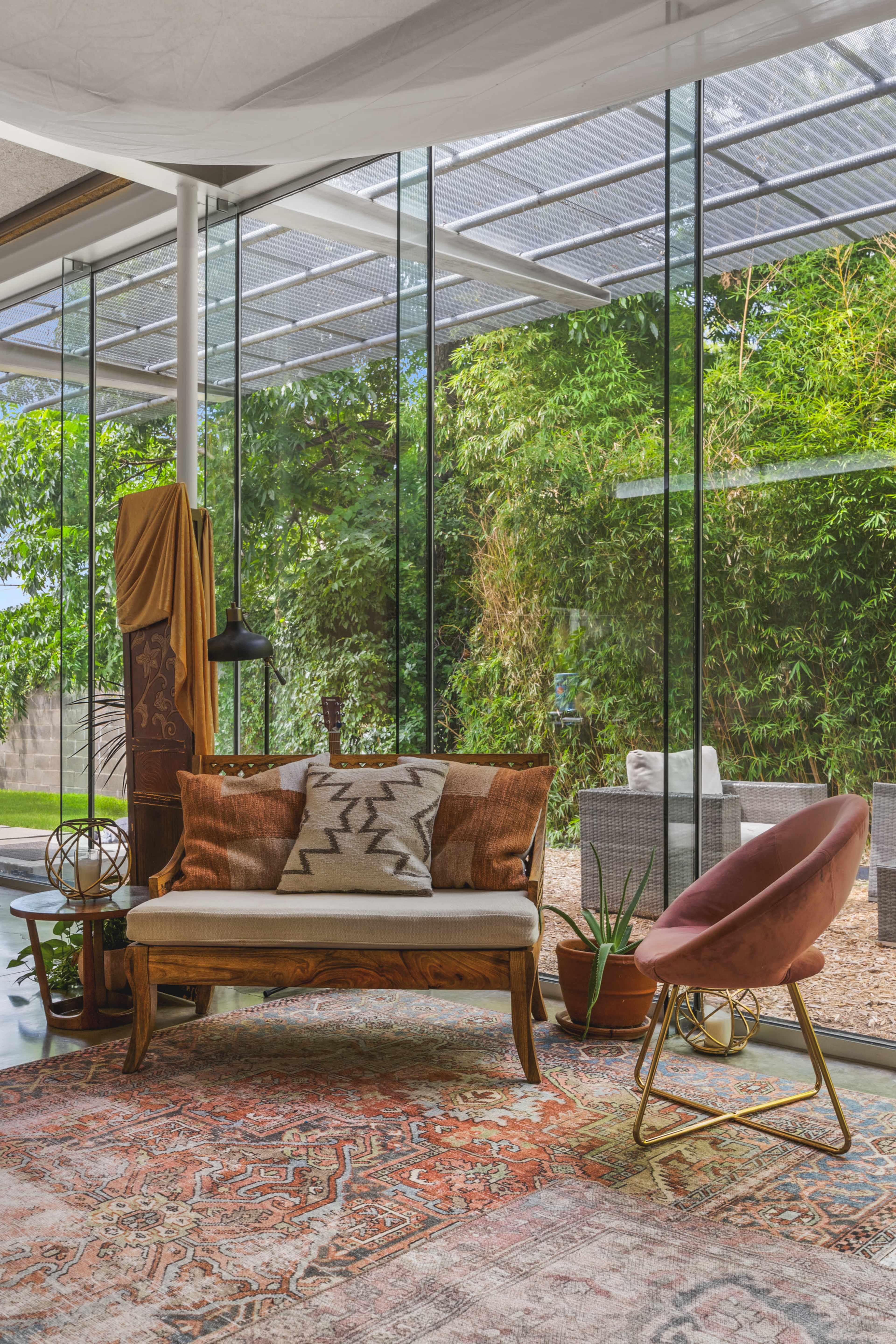 A cozy sitting area features a wooden bench with decorative pillows, a pink chair, and plants, surrounded by large glass windows overlooking a green garden.