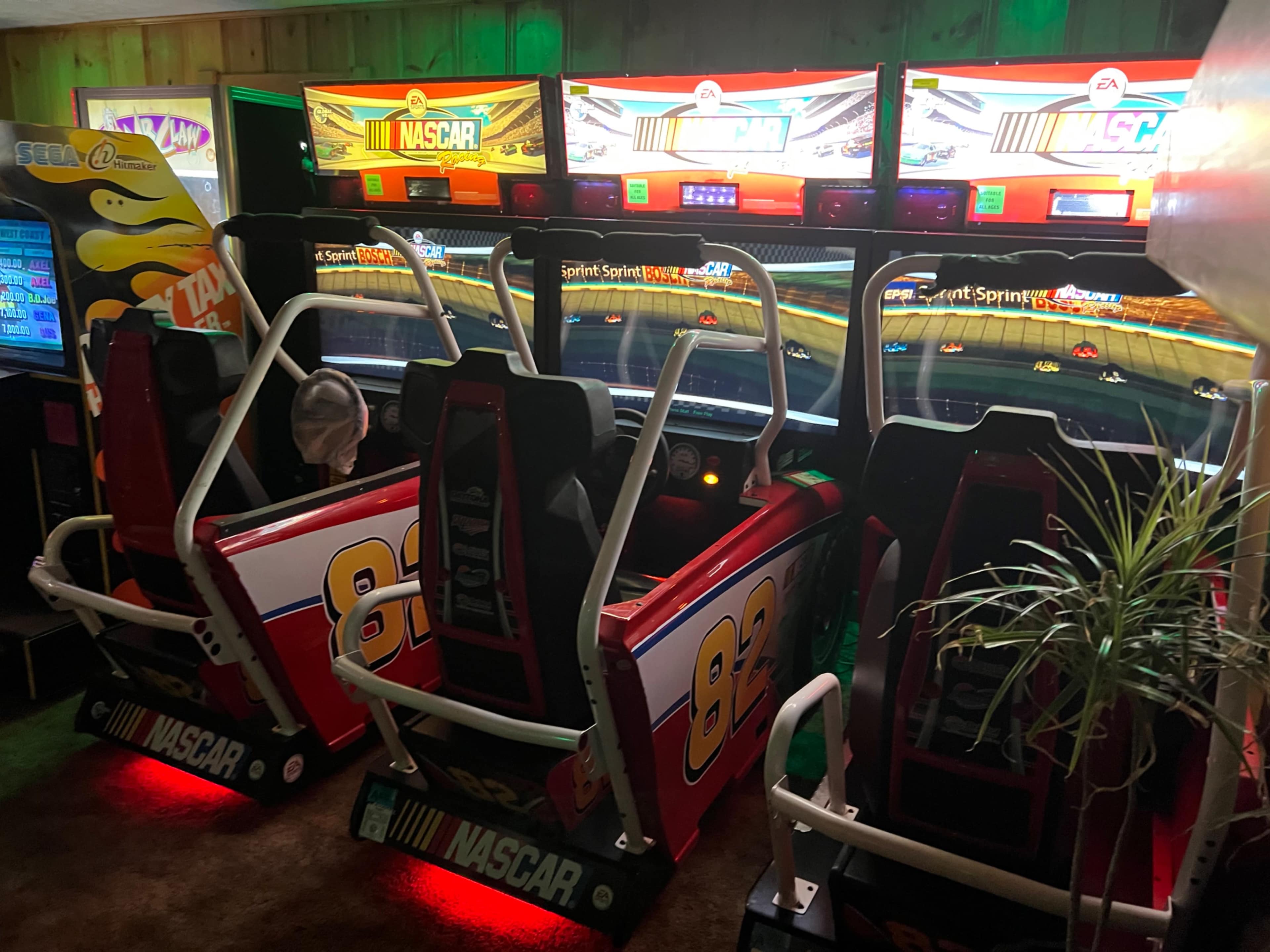 The image shows three NASCAR-themed arcade racing machines lined up in a dimly lit room.