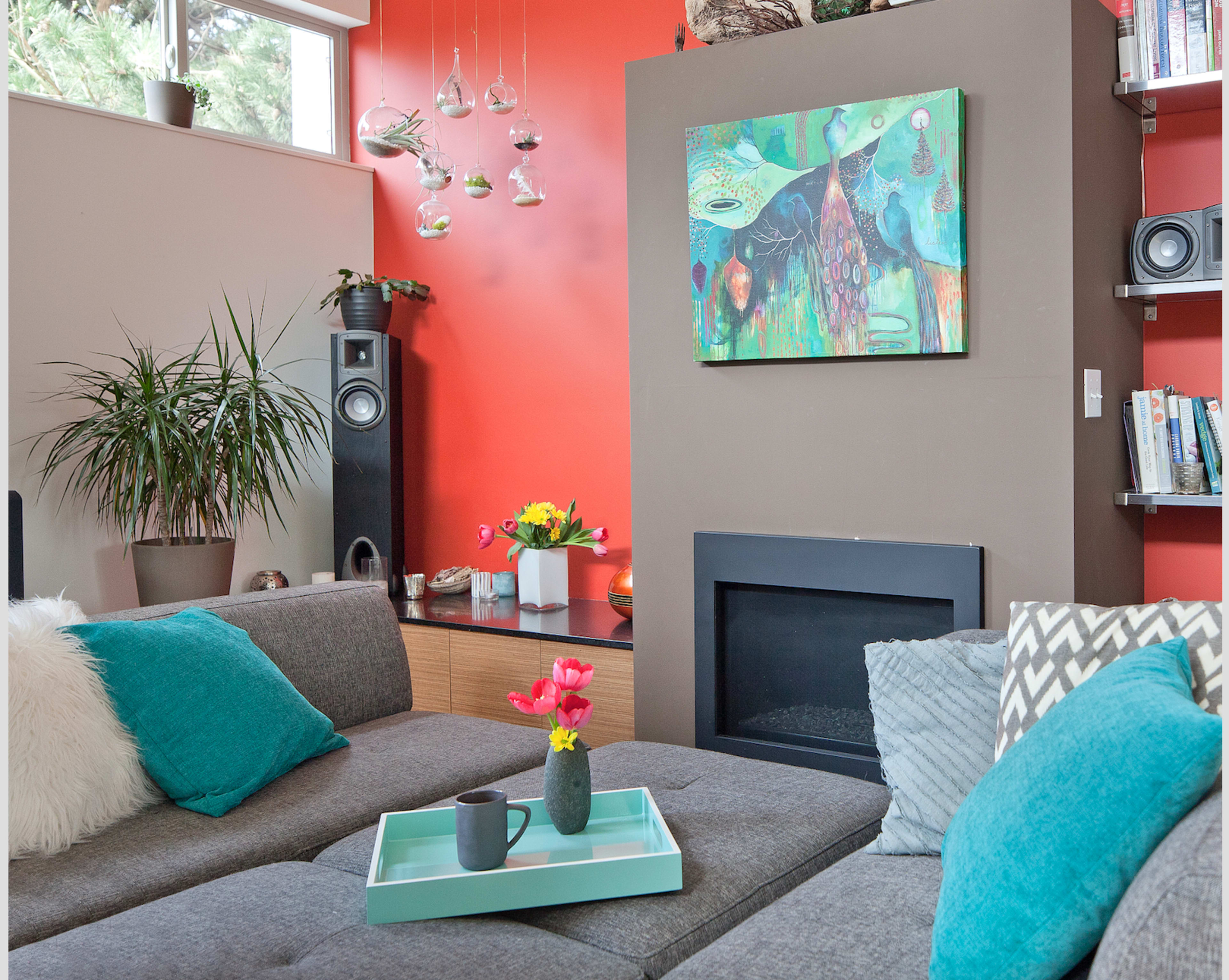 A cozy living room featuring a gray sectional sofa with teal cushions, a modern fireplace, and vibrant red walls adorned with hanging planters and a colorful painting.