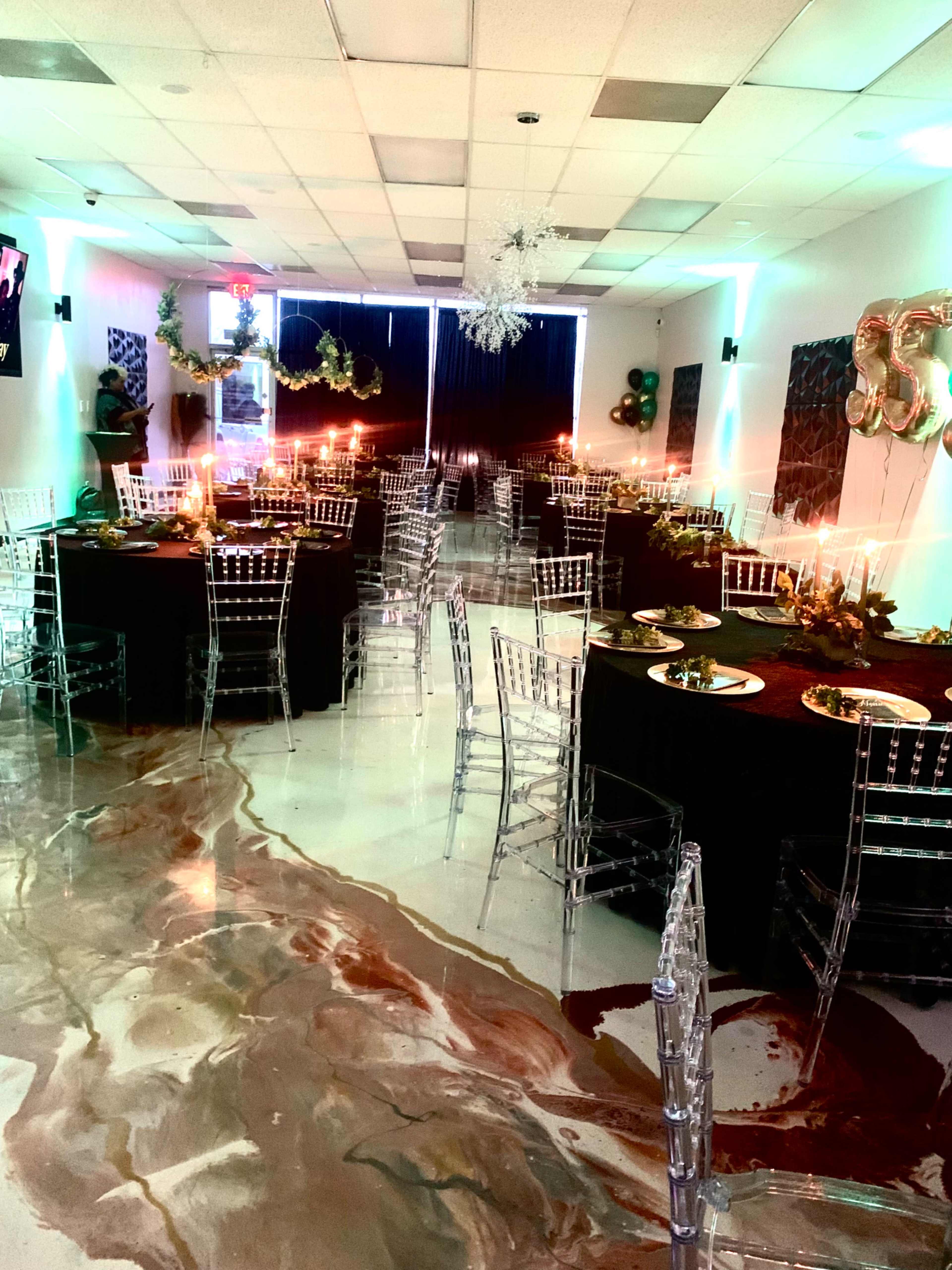 A dining area set up for an event, featuring black tablecloths, clear chiavari chairs, and decorative centerpieces with candles on elegantly arranged tables.