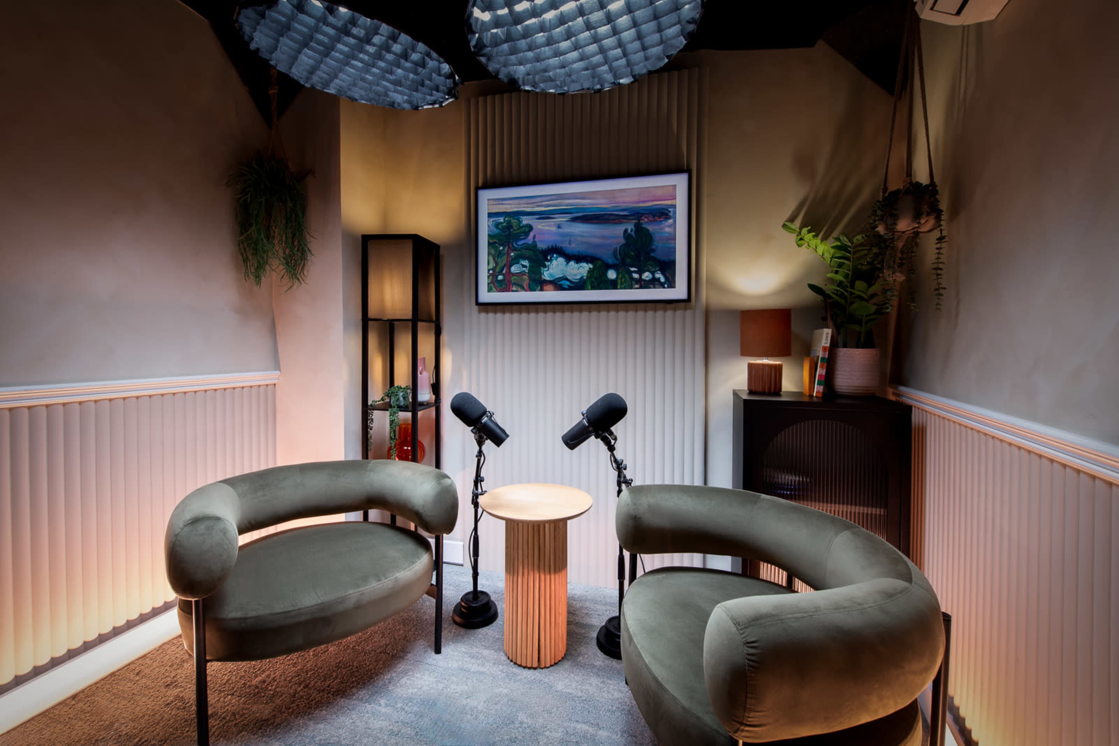 The image depicts a small, modern recording studio featuring two curved chairs, a round table between them, and two microphones positioned in front of the chairs, with a framed painting on the wall.