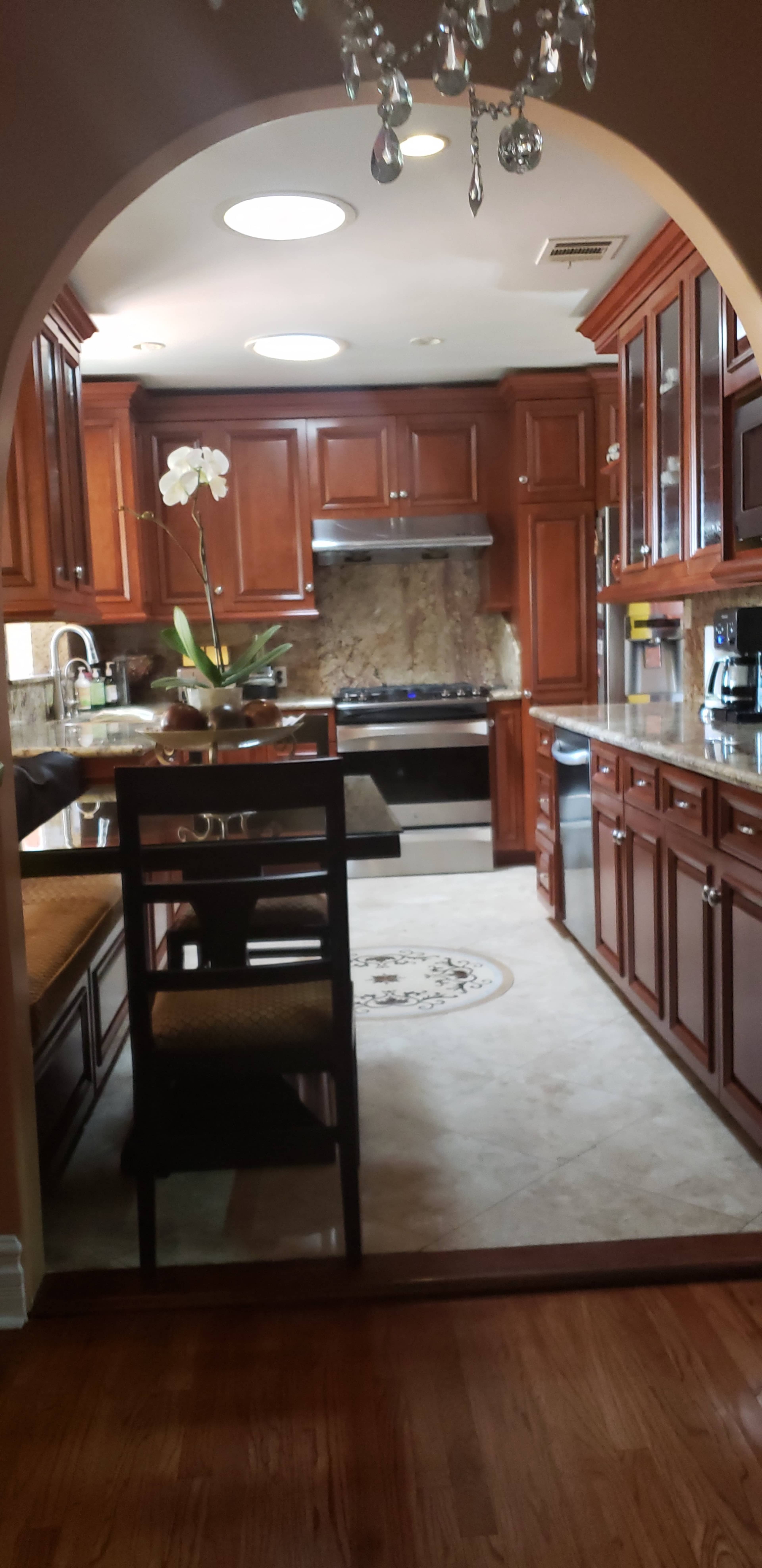 The photo shows a wooden kitchen with dark cabinets, a central island, and stainless steel appliances, viewed through an arched entryway.
