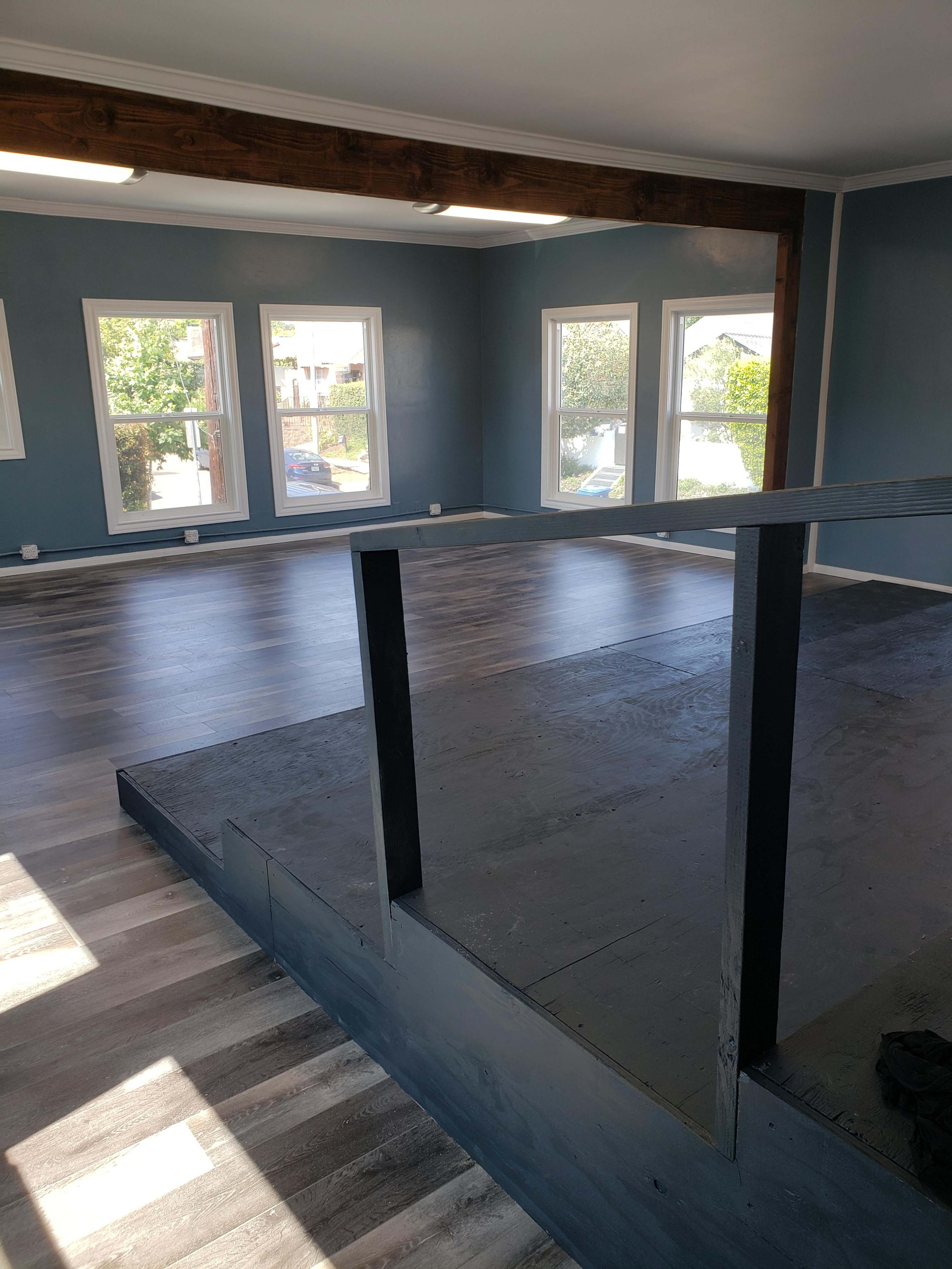 An empty room with a wooden beam across the ceiling, large windows allowing natural light, and a raised platform on the floor.