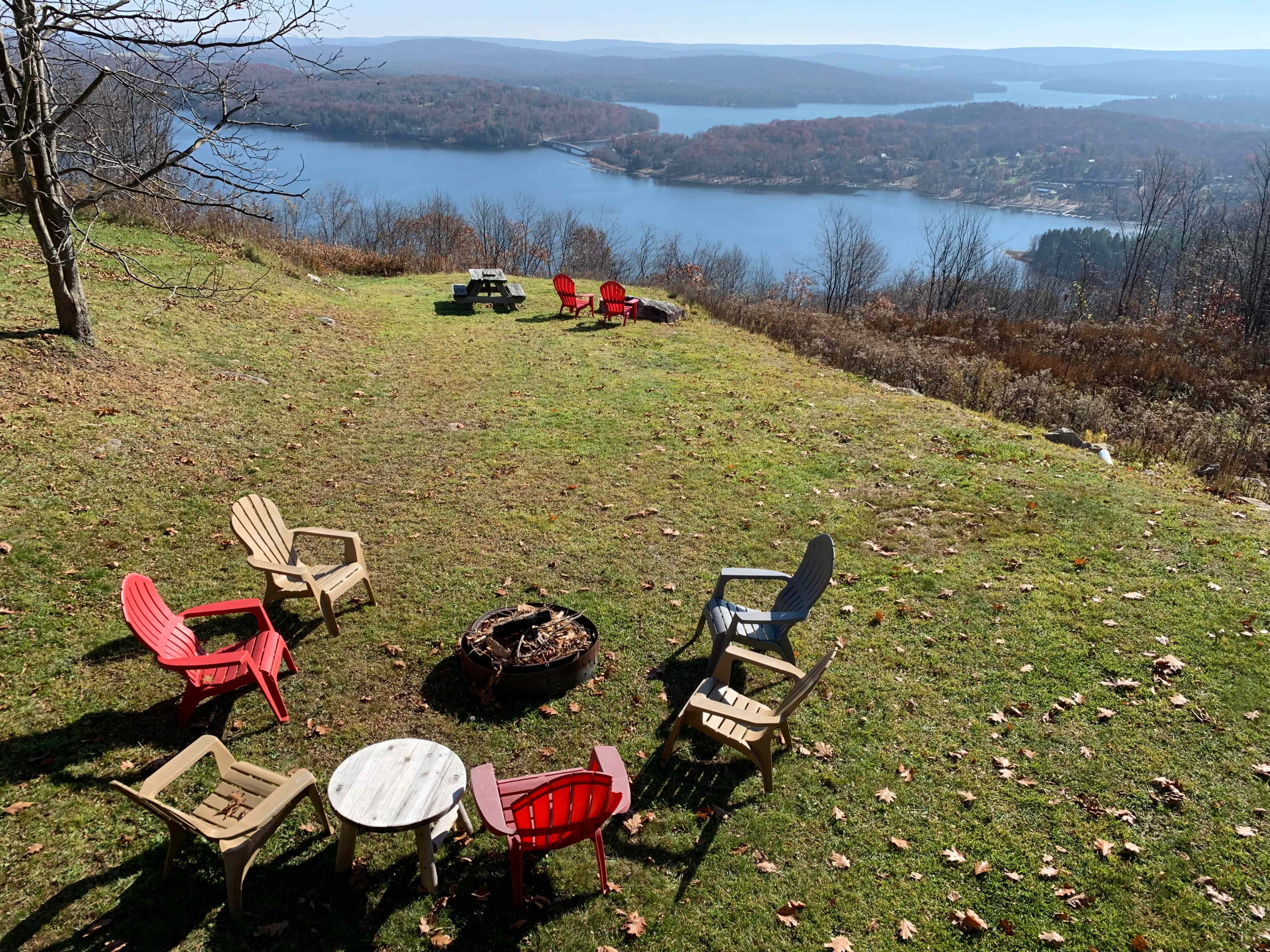 A circle of outdoor chairs surrounds a fire pit on a grassy slope overlooking a lake and forested hills.
