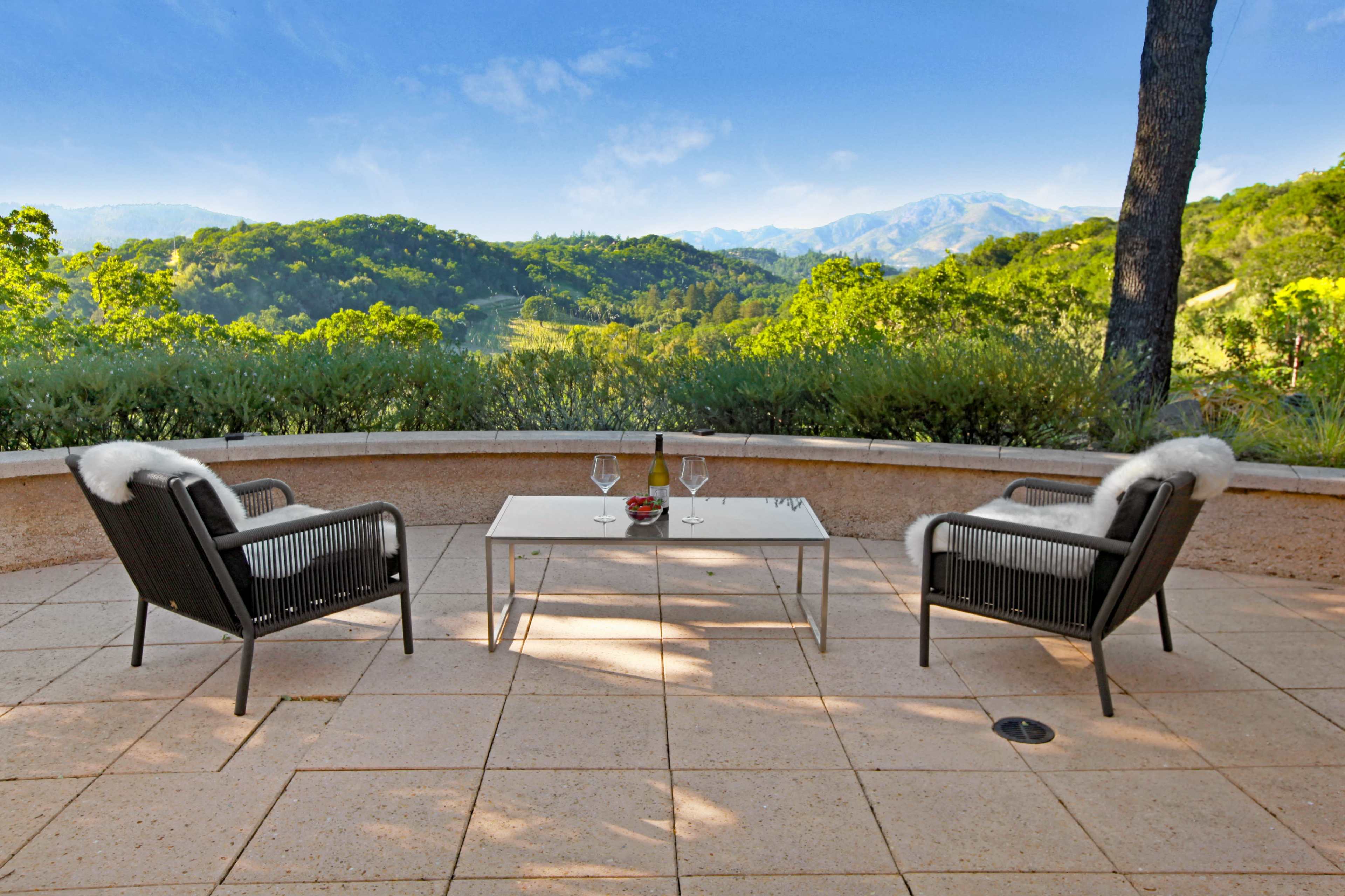 Two chairs with white fur cushions are positioned around a glass table, overlooking a scenic view of rolling hills and mountains.