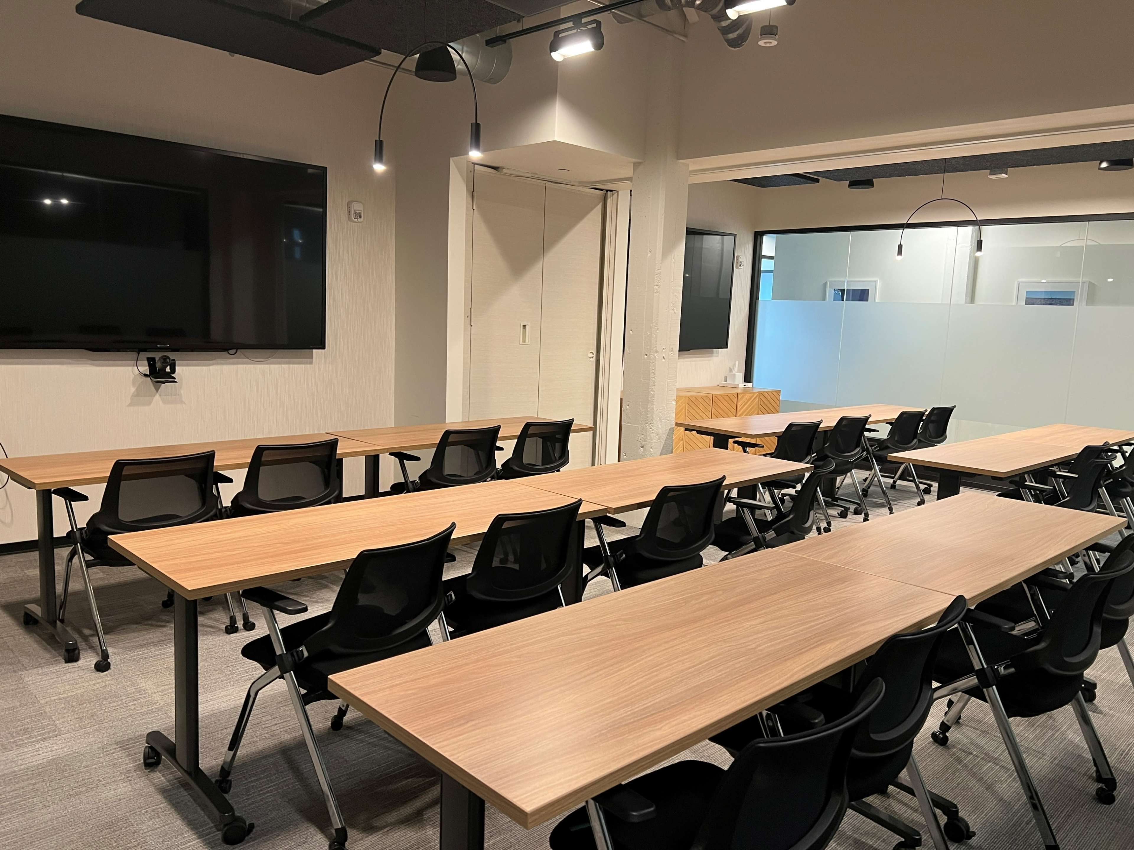 A modern conference room features multiple rows of wooden tables and black chairs, with large screens on the walls.