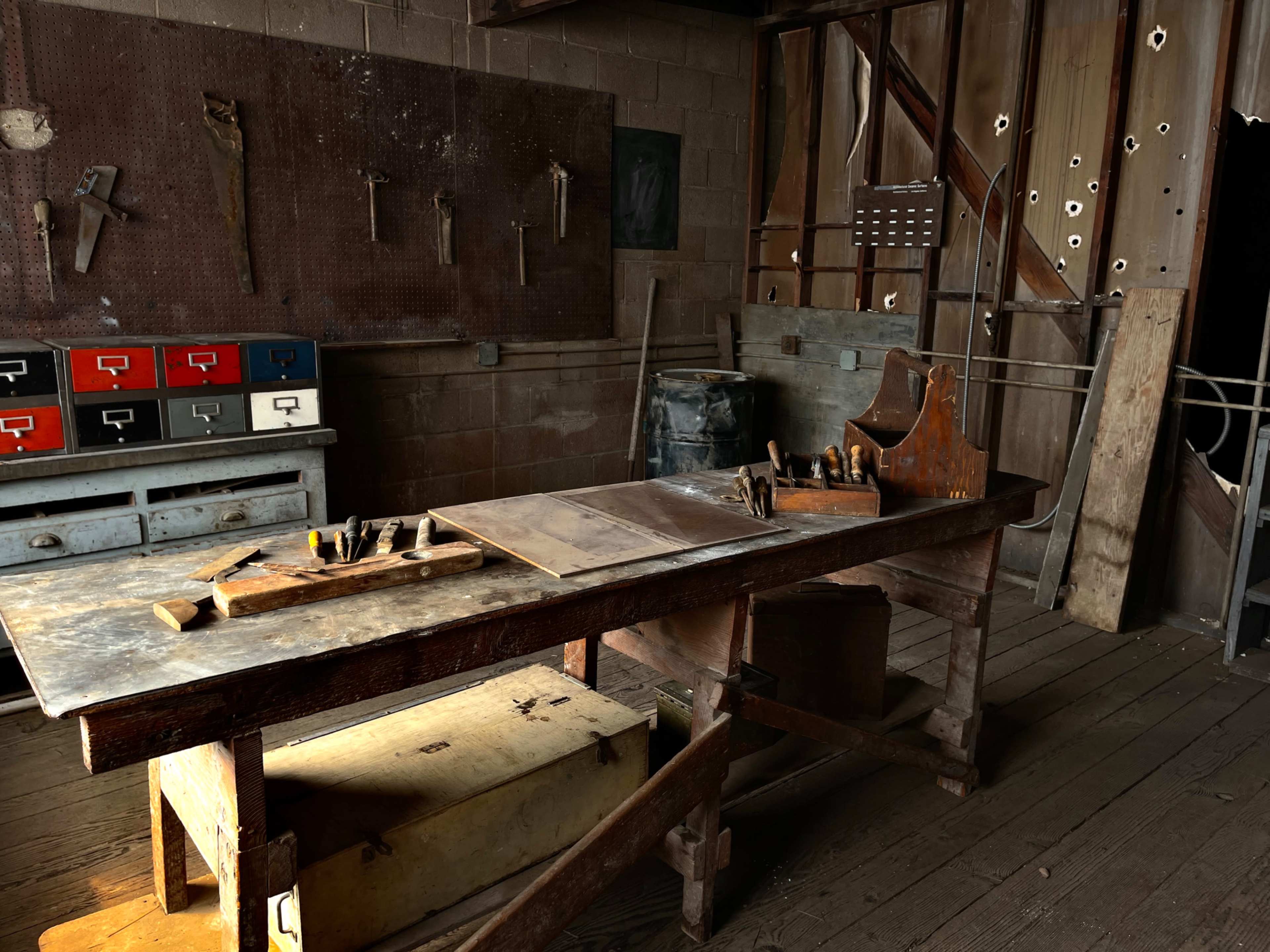 A woodworking shop features a wooden table with tools, surrounded by storage drawers and a partially exposed wall.