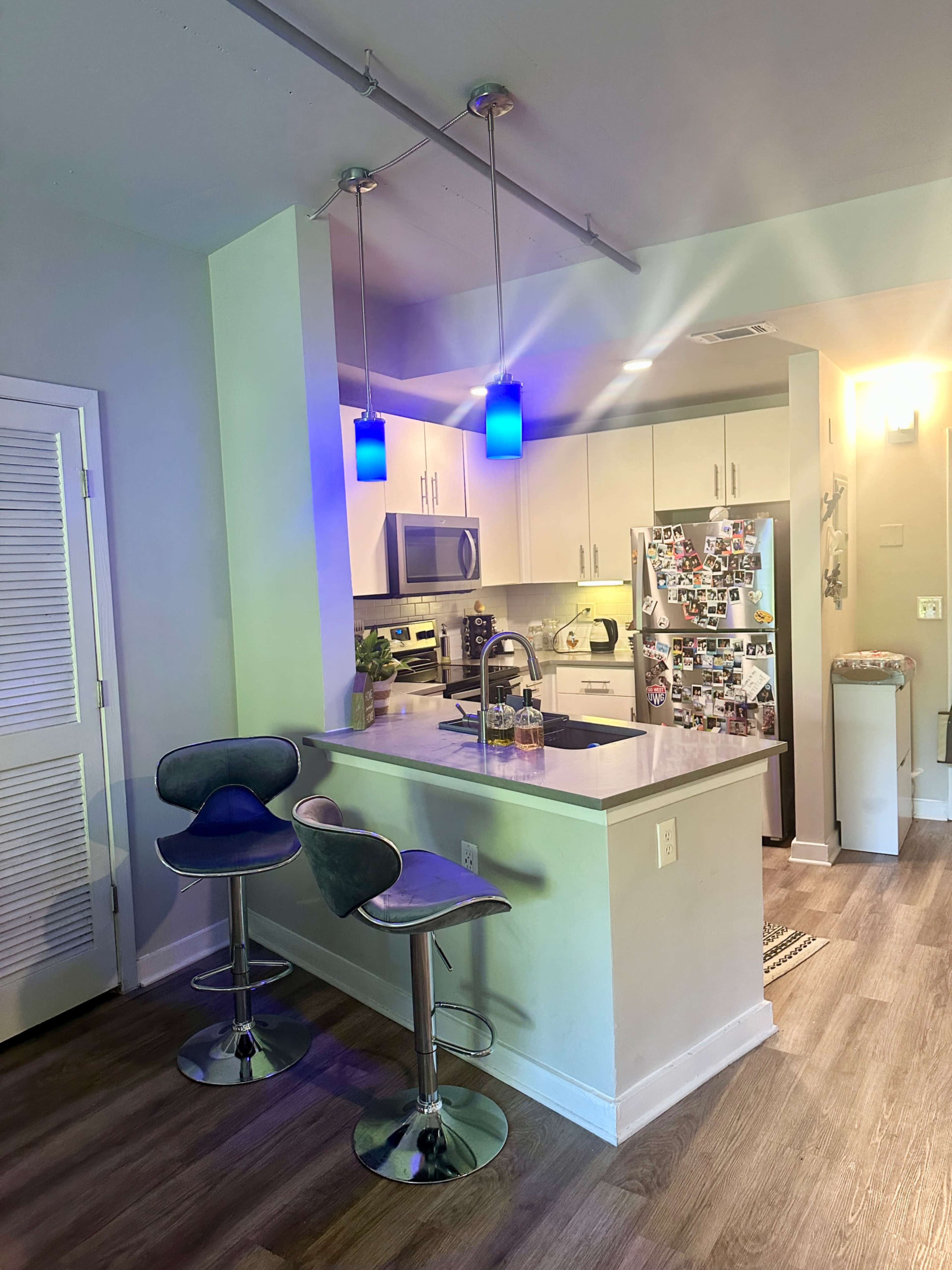 The image shows a modern kitchen with a central island, two bar stools, pendant lighting, and stainless steel appliances.