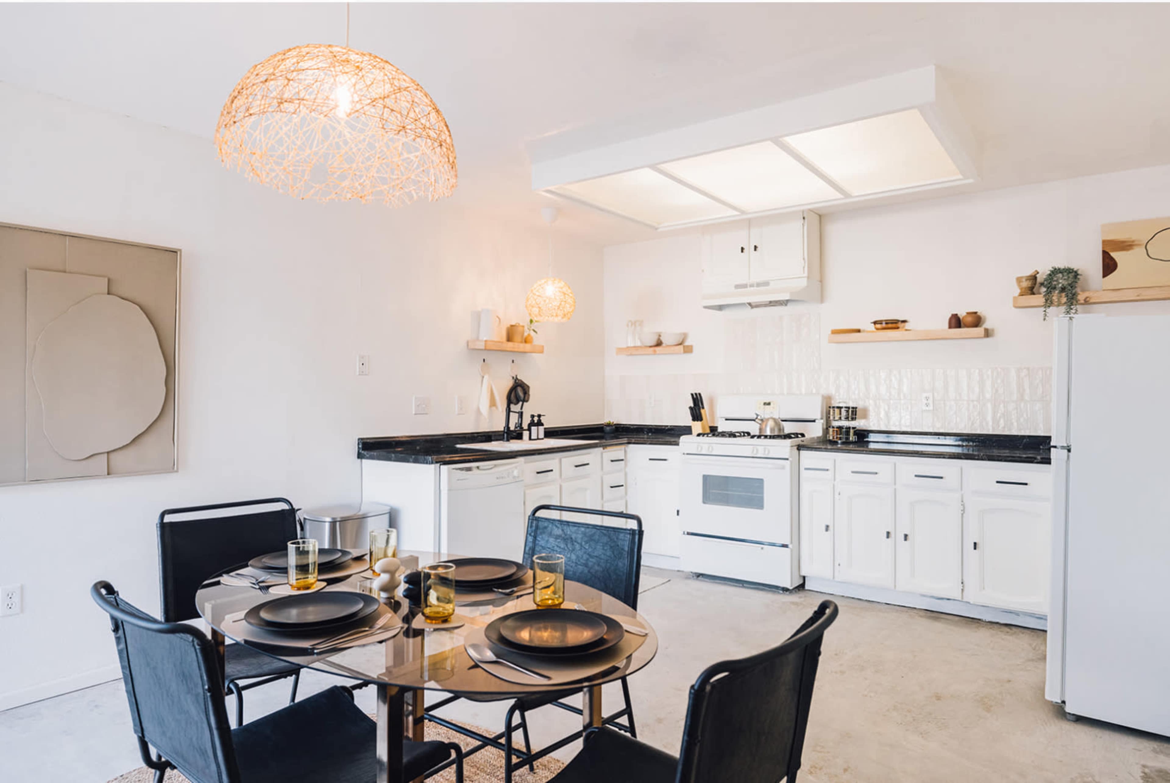 A minimalistic kitchen and dining area features a round table set for four, equipped with black chairs, a light fixture, and a white cabinetry backdrop.