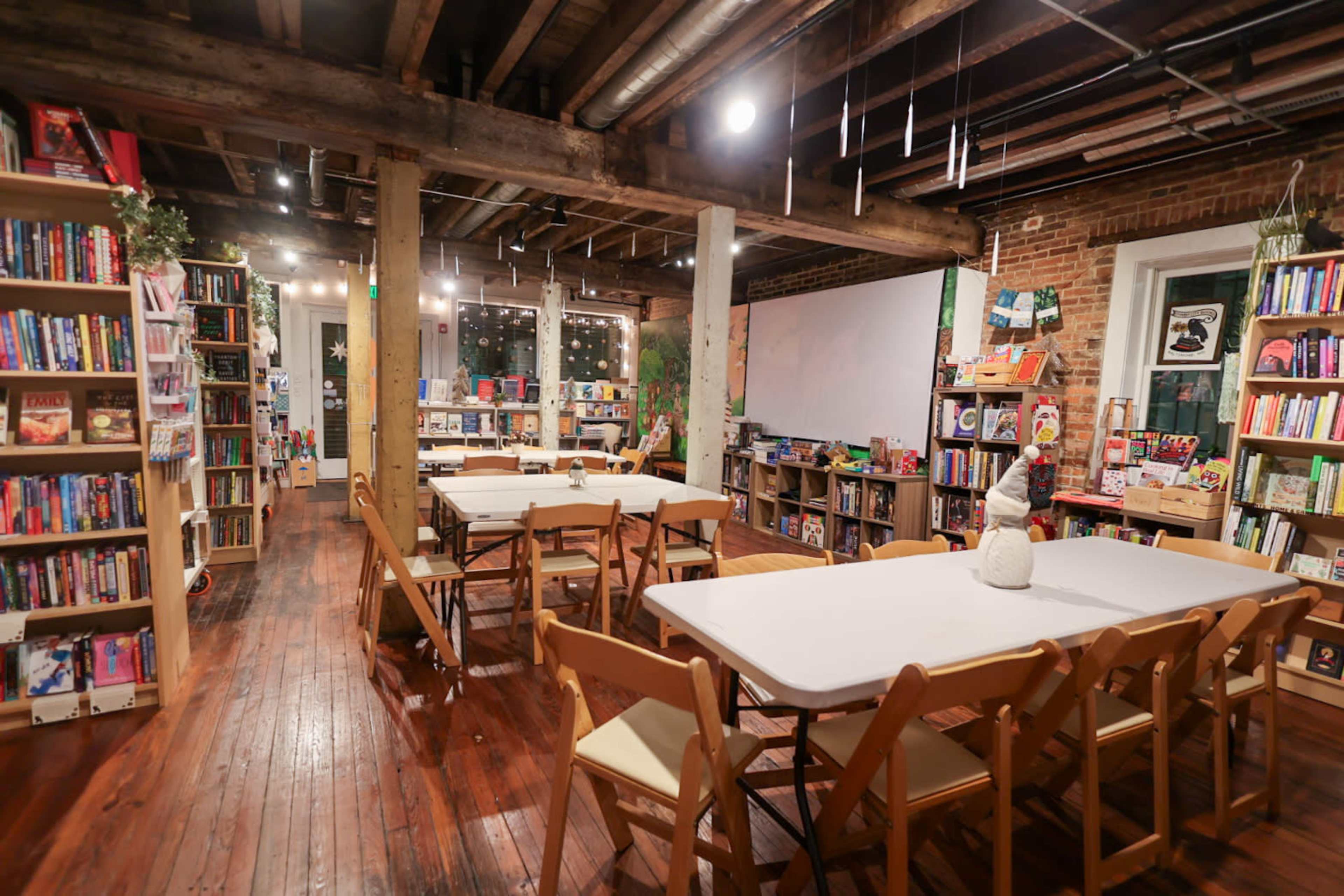 The image shows a cozy bookstore with wooden beams, shelves filled with books, and tables set up for gatherings or events.