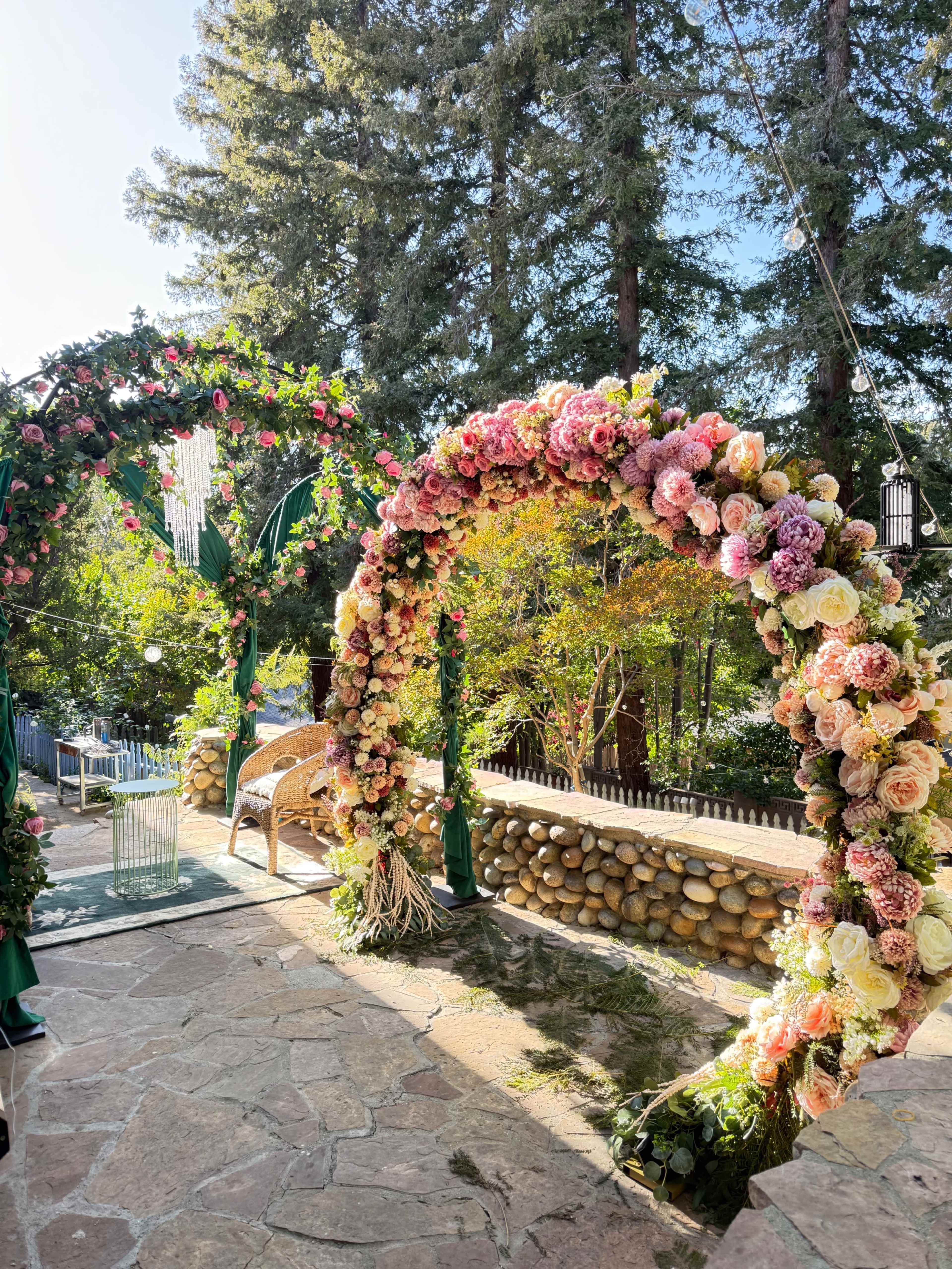 The scene features two ornate floral arches, one adorned with pink and white blooms, set against a backdrop of tall trees and a stone pathway.