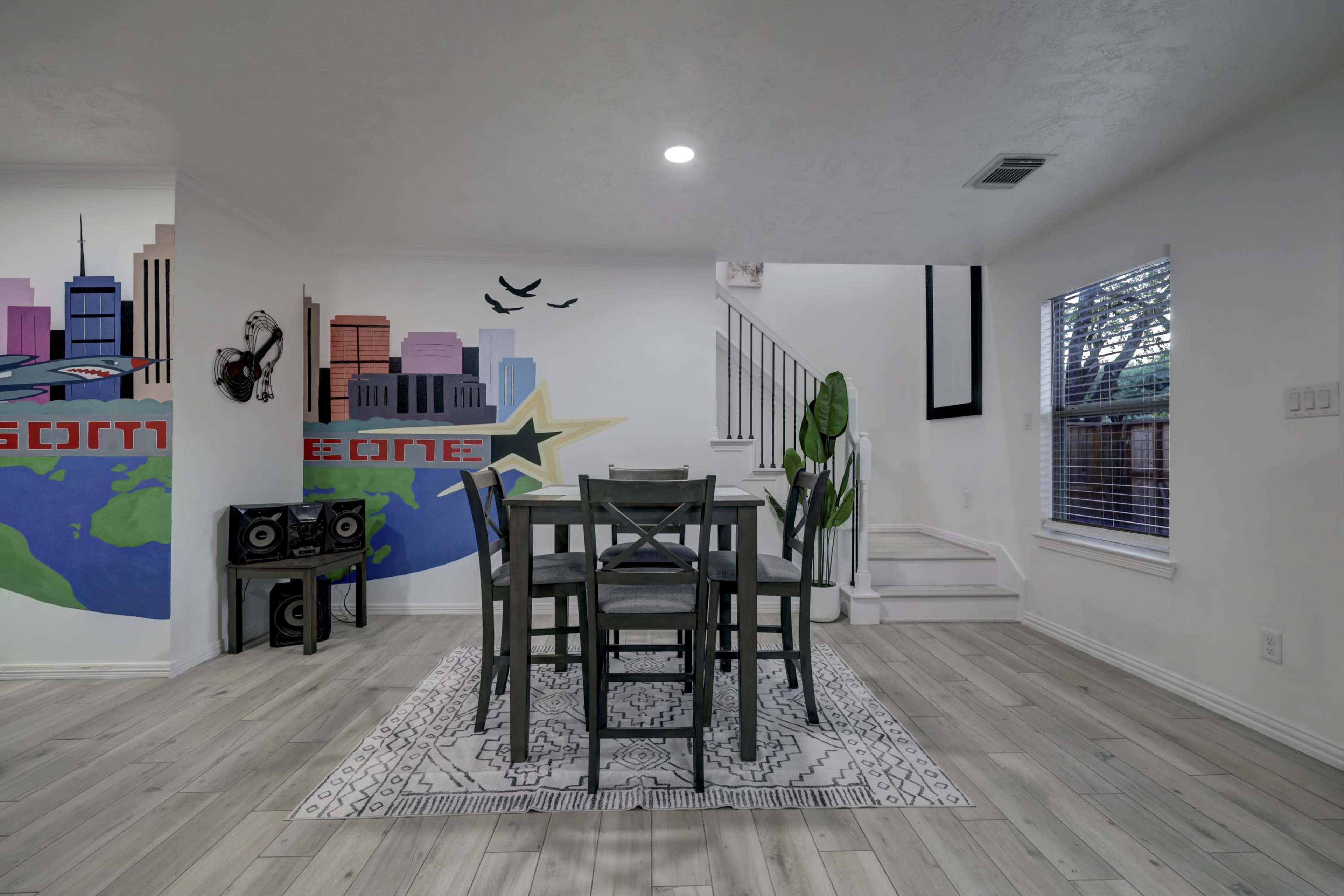 A dining area with a table and chairs, featuring a colorful wall mural of a city skyline and a staircase in the background.