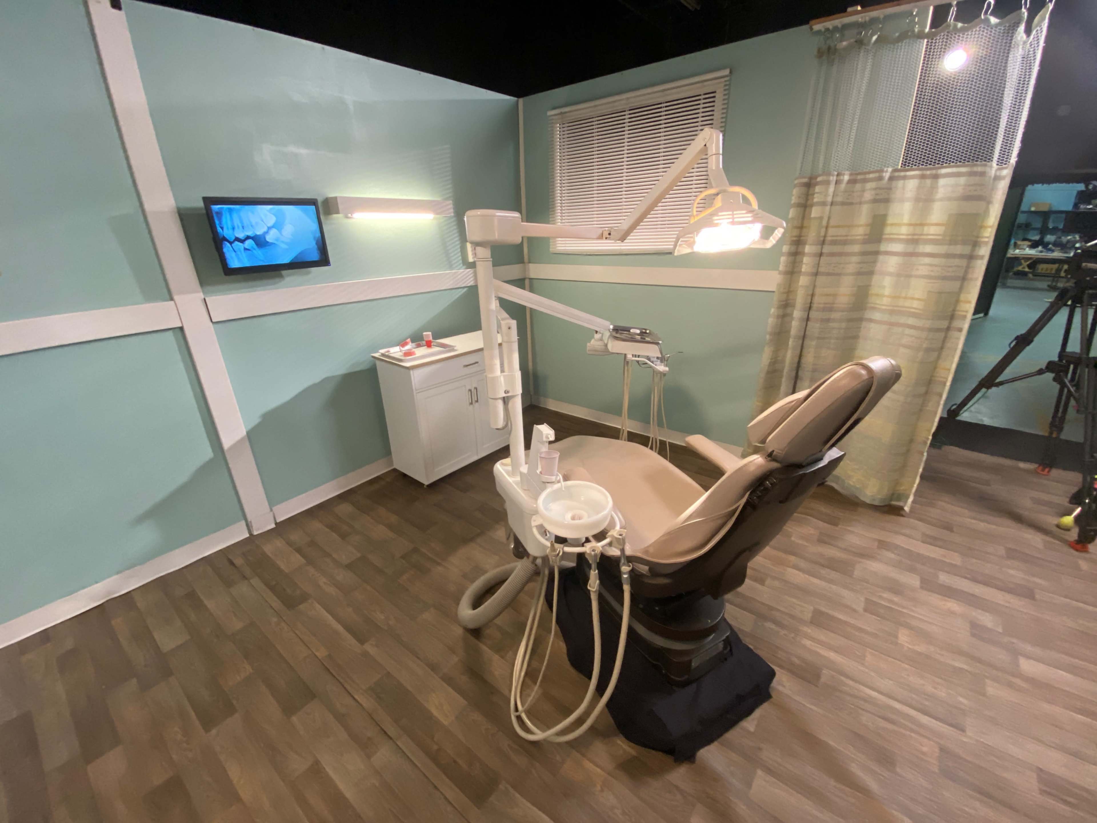 The image shows a dental exam room equipped with a dental chair, monitor, and a small storage cabinet against a light green wall.