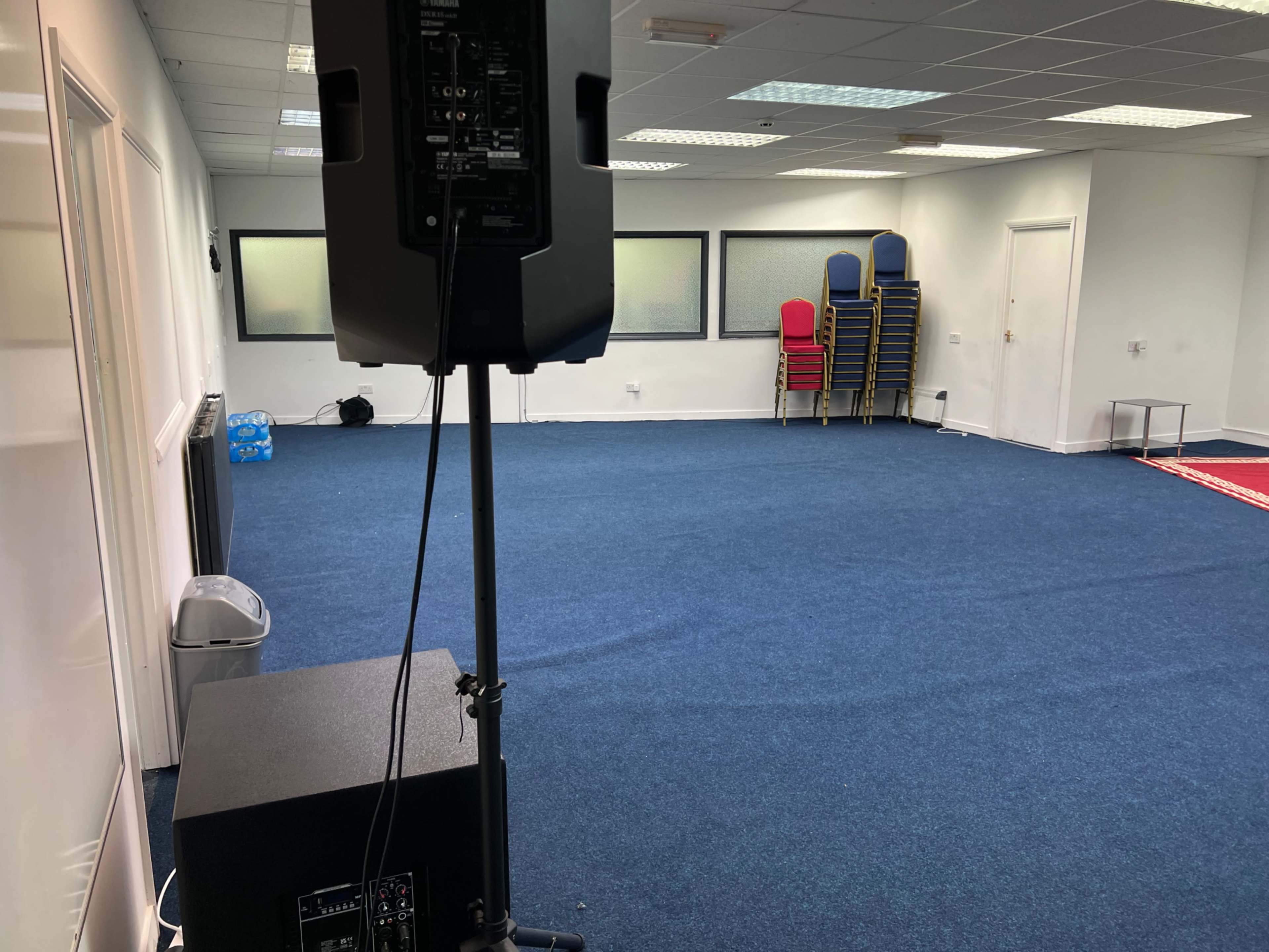 The image shows a spacious, empty room with blue carpet, a speaker on a stand, and stacked chairs along one wall.