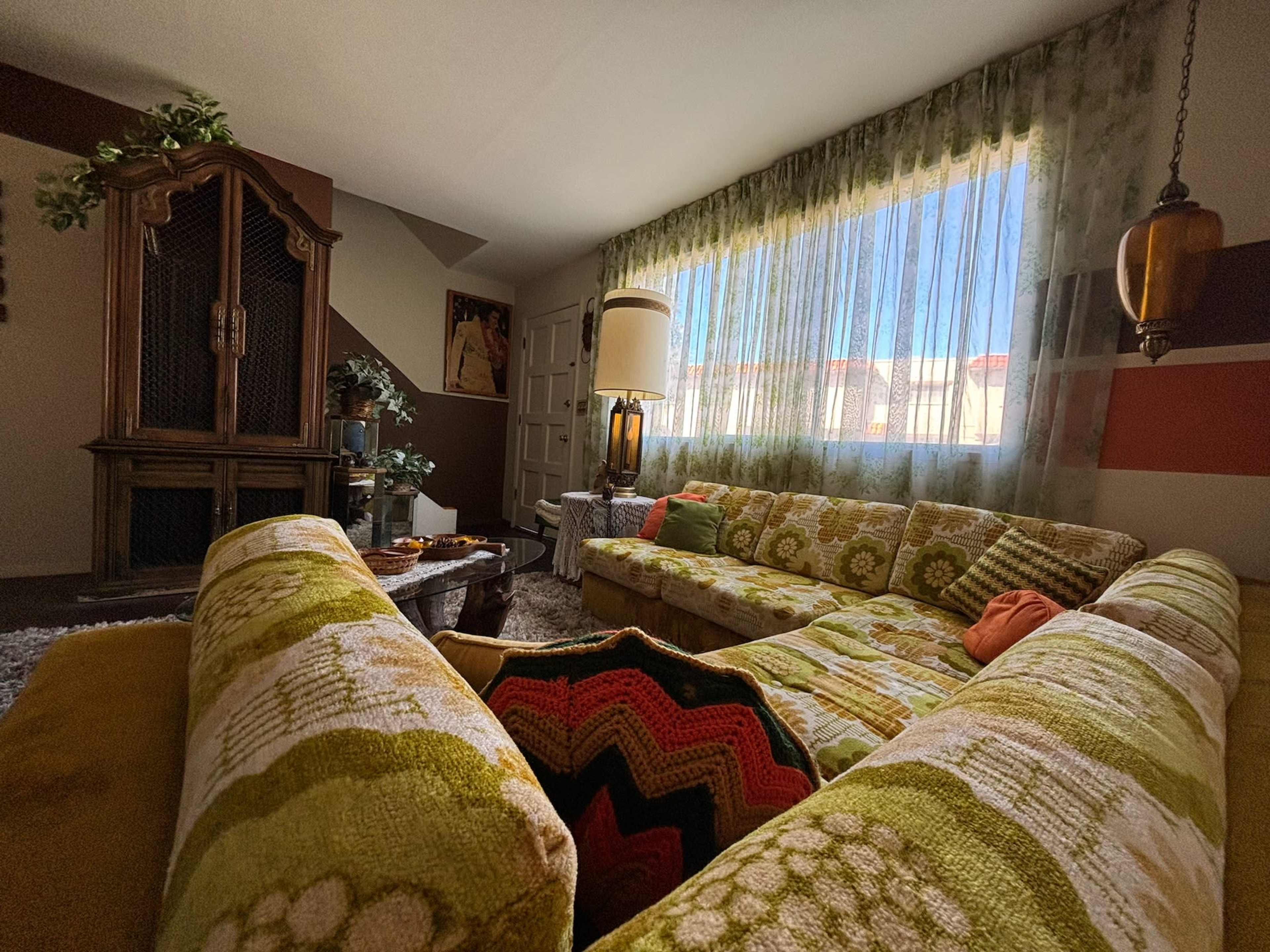The image shows a living room featuring a large, patterned green sectional sofa, a vintage wooden cabinet, and a window with sheer curtains allowing sunlight to enter.