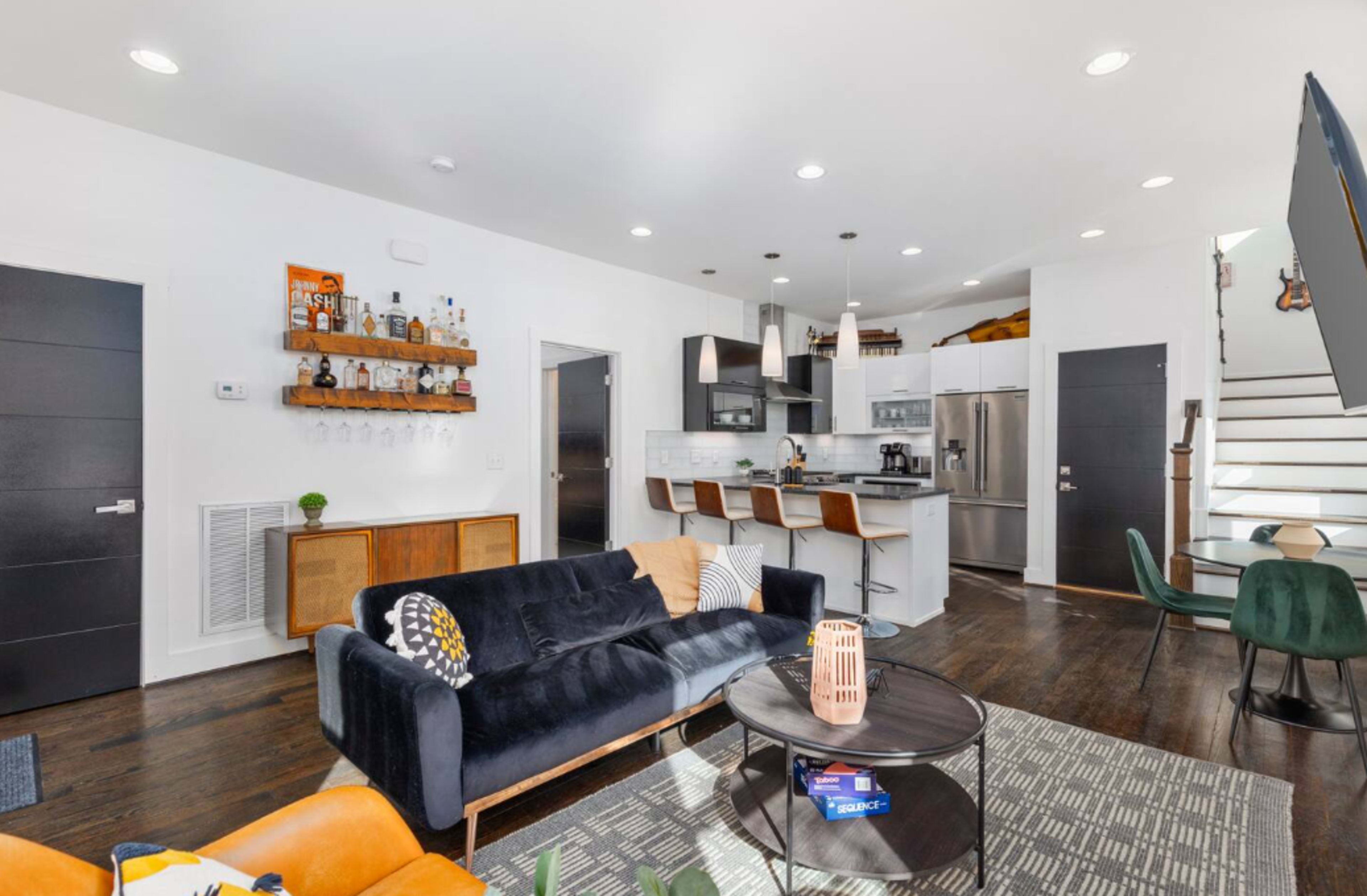 A modern living room with a black sofa, a round coffee table, bar stools at a kitchen counter, and a staircase leading to the upper level.