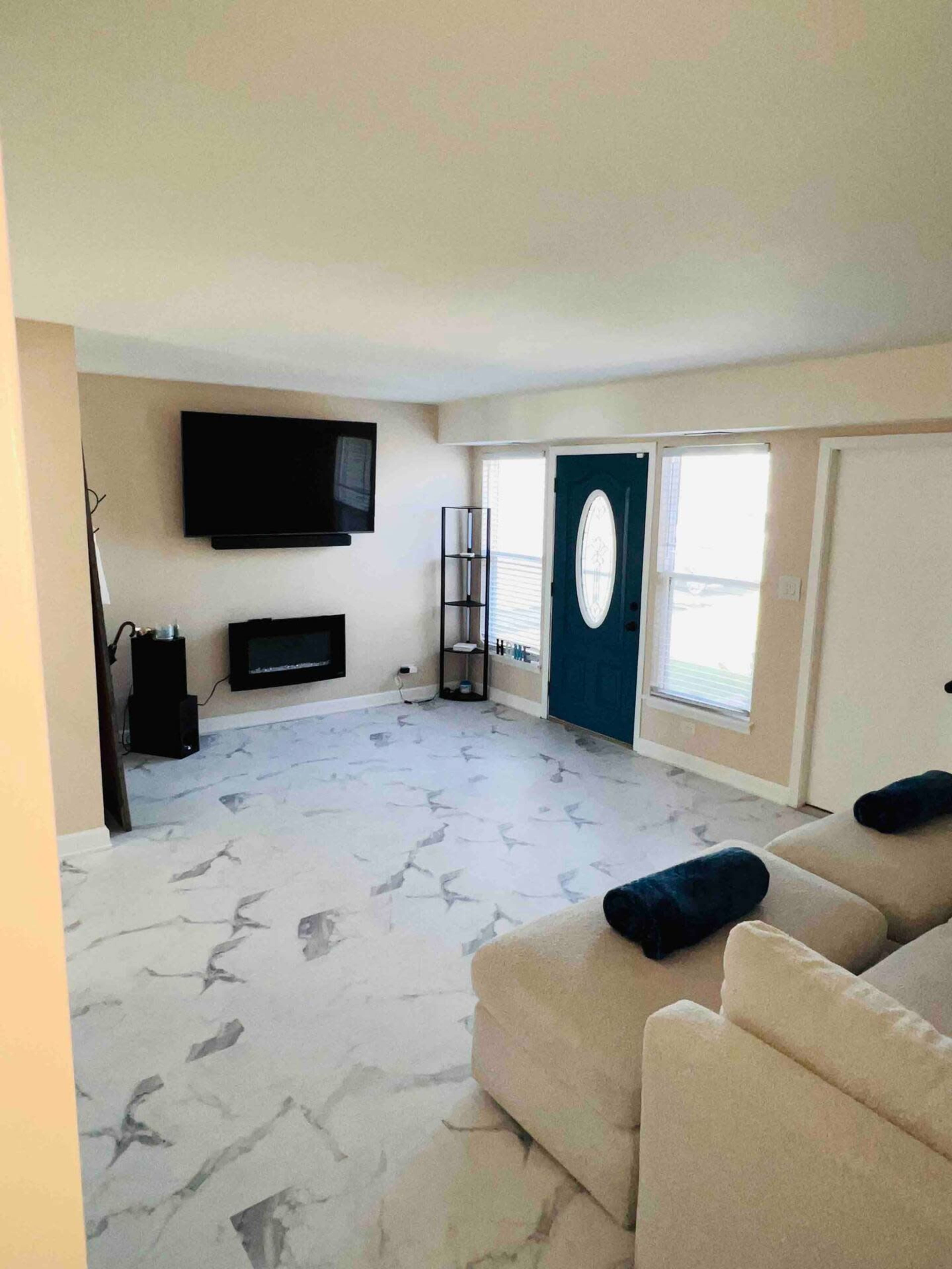 A living room features a television mounted on the wall, a modern electric fireplace, a blue front door, large windows, and a light-colored sectional sofa with dark throw pillows.