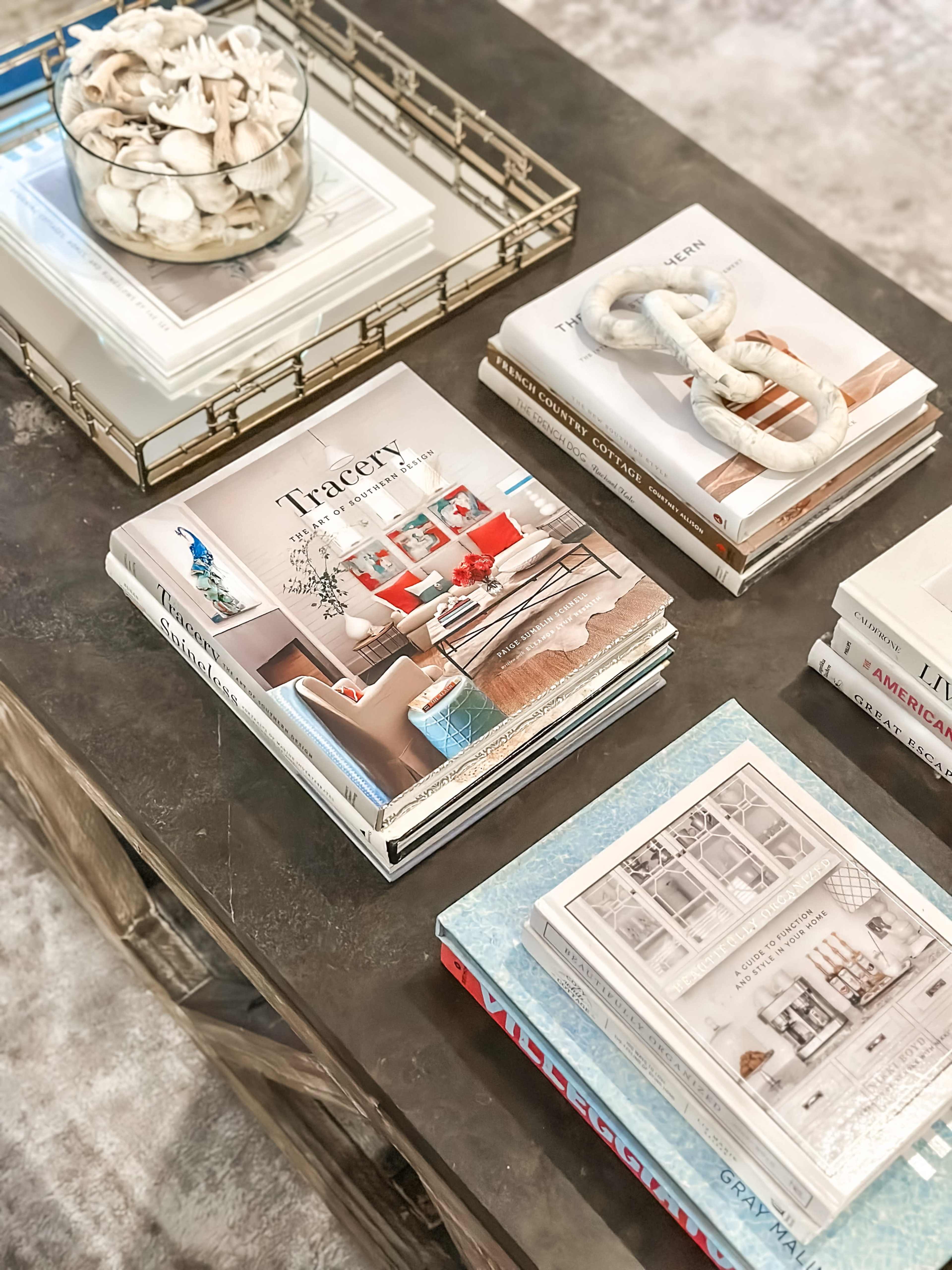 The image shows a coffee table arrangement featuring several stacked books and a decorative bowl filled with shells.