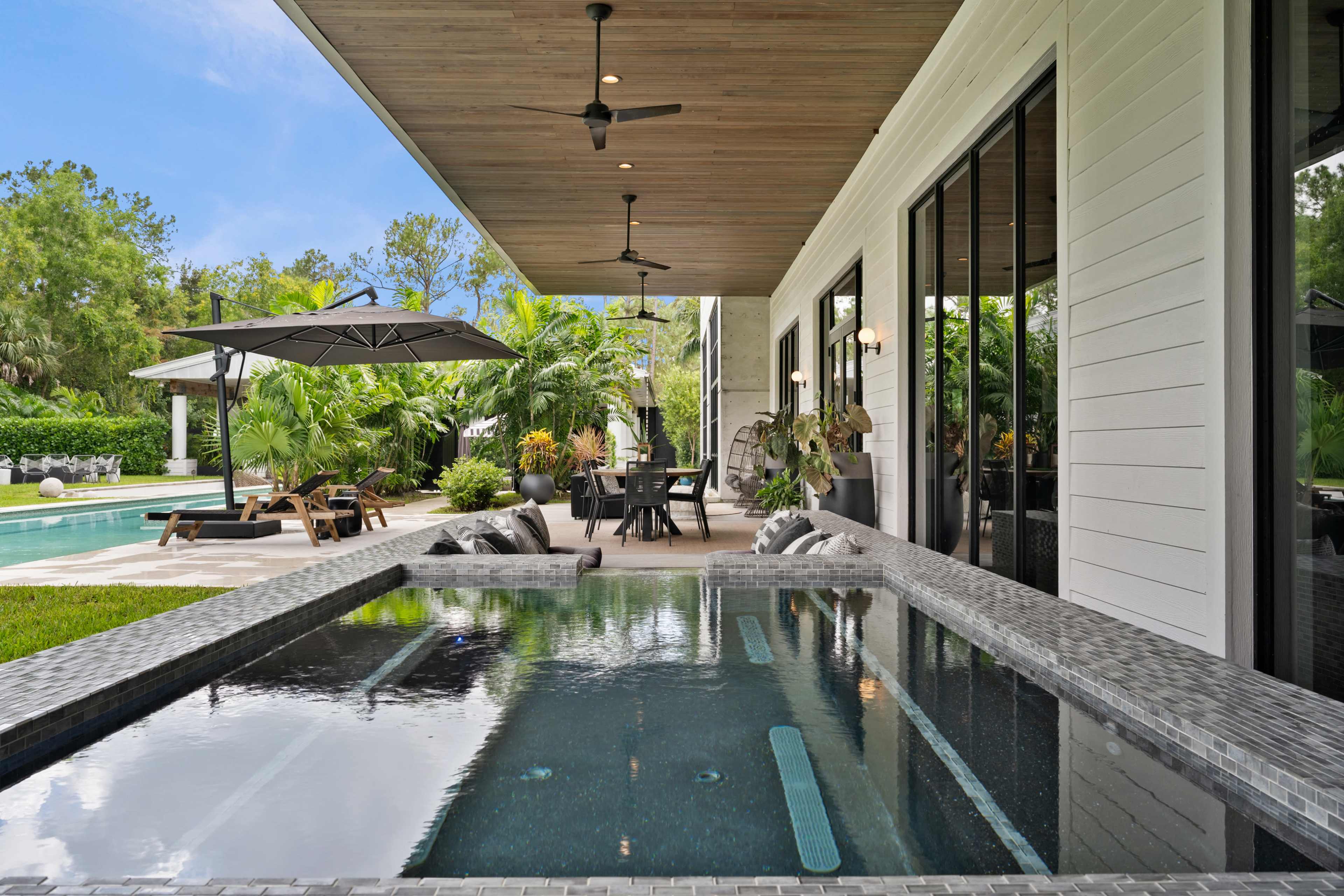 The image shows a modern outdoor space featuring a shallow pool with a spa area, surrounded by lush greenery and a dining area under an umbrella.