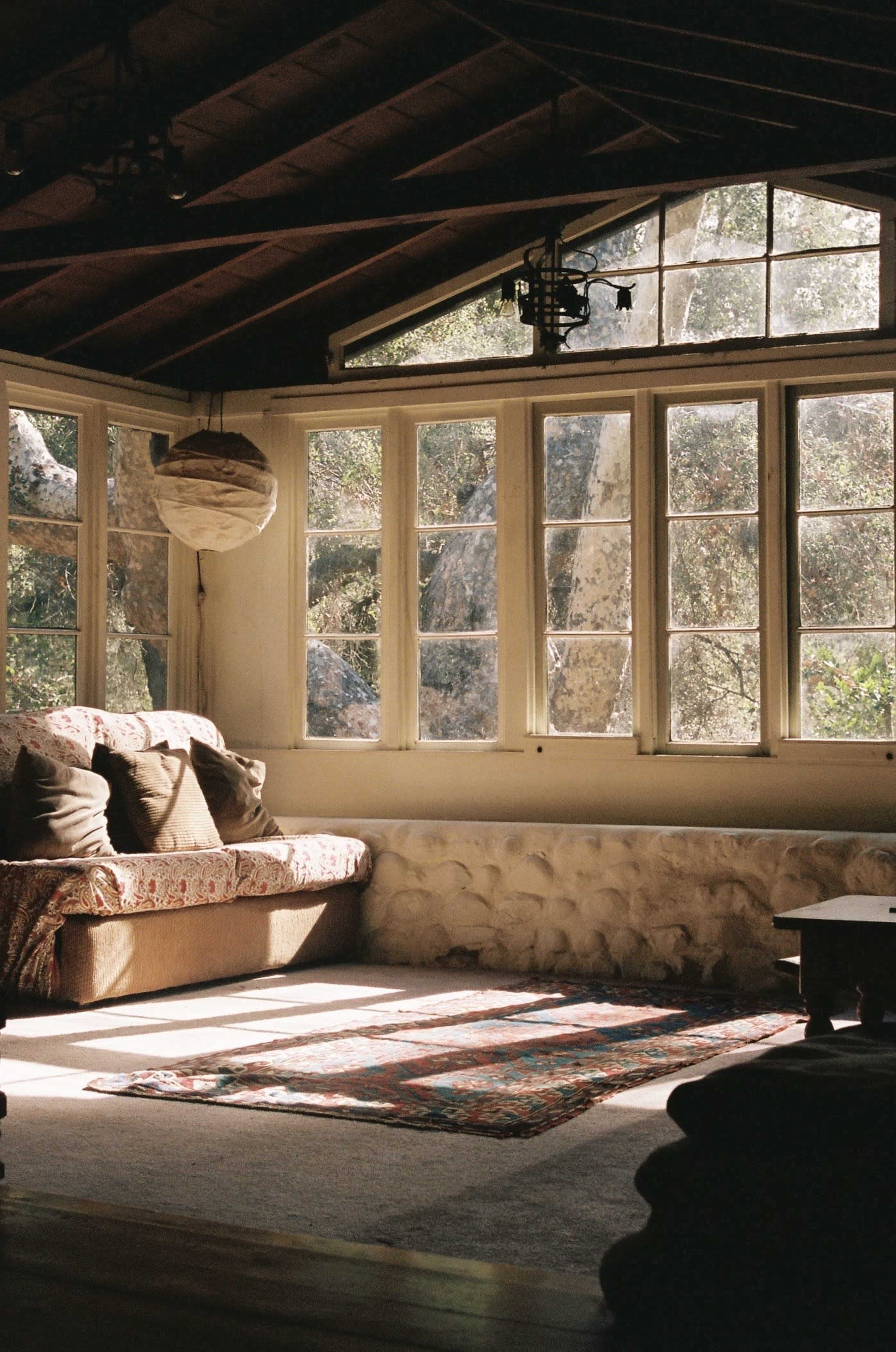 A cozy living room features a sofa, large windows letting in natural light, and a patchwork rug on the wooden floor.