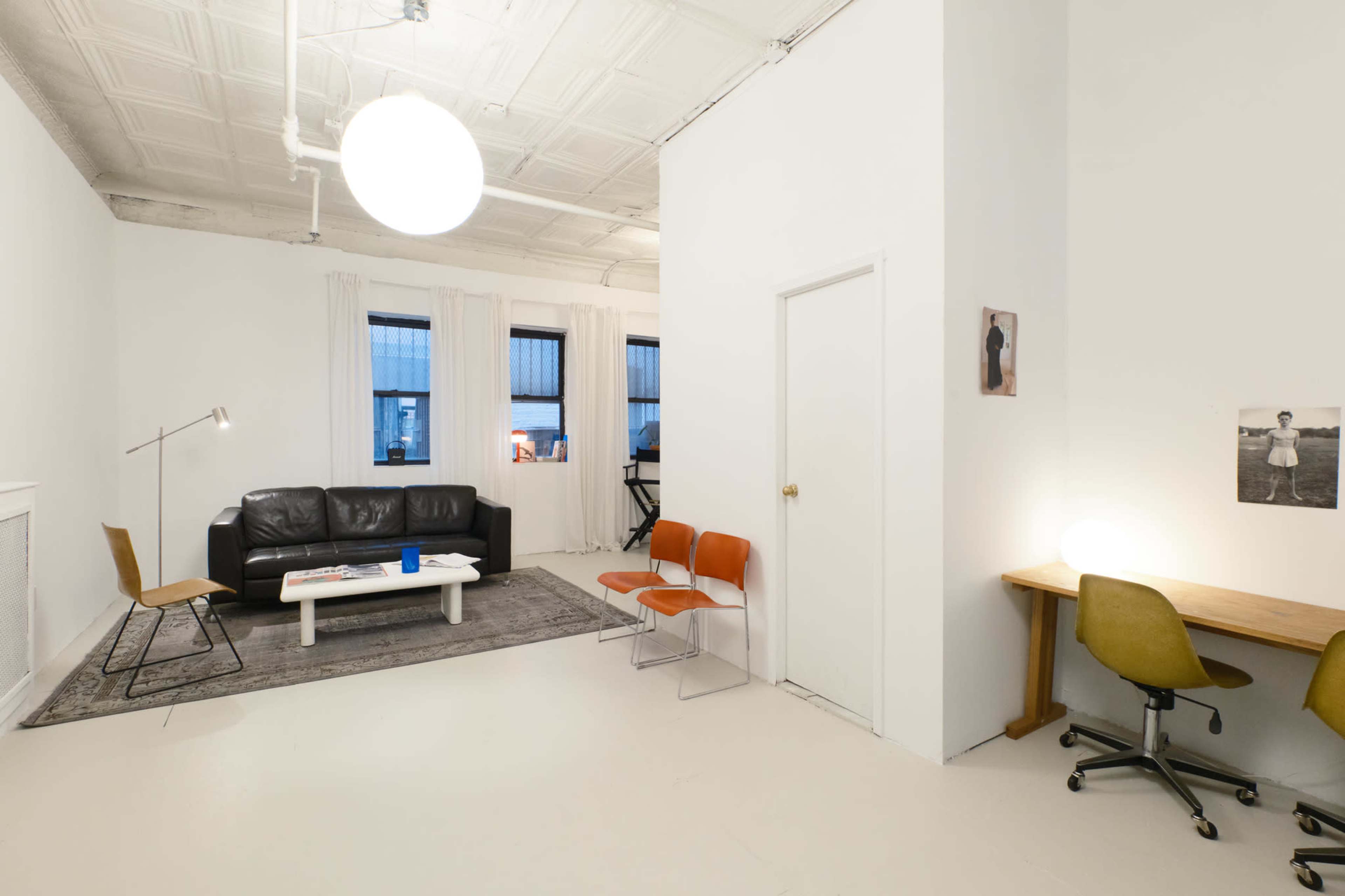 The image shows a minimalist living room featuring a black leather couch, a coffee table, a gray rug, and a small desk with a chair, all set against white walls and large windows.