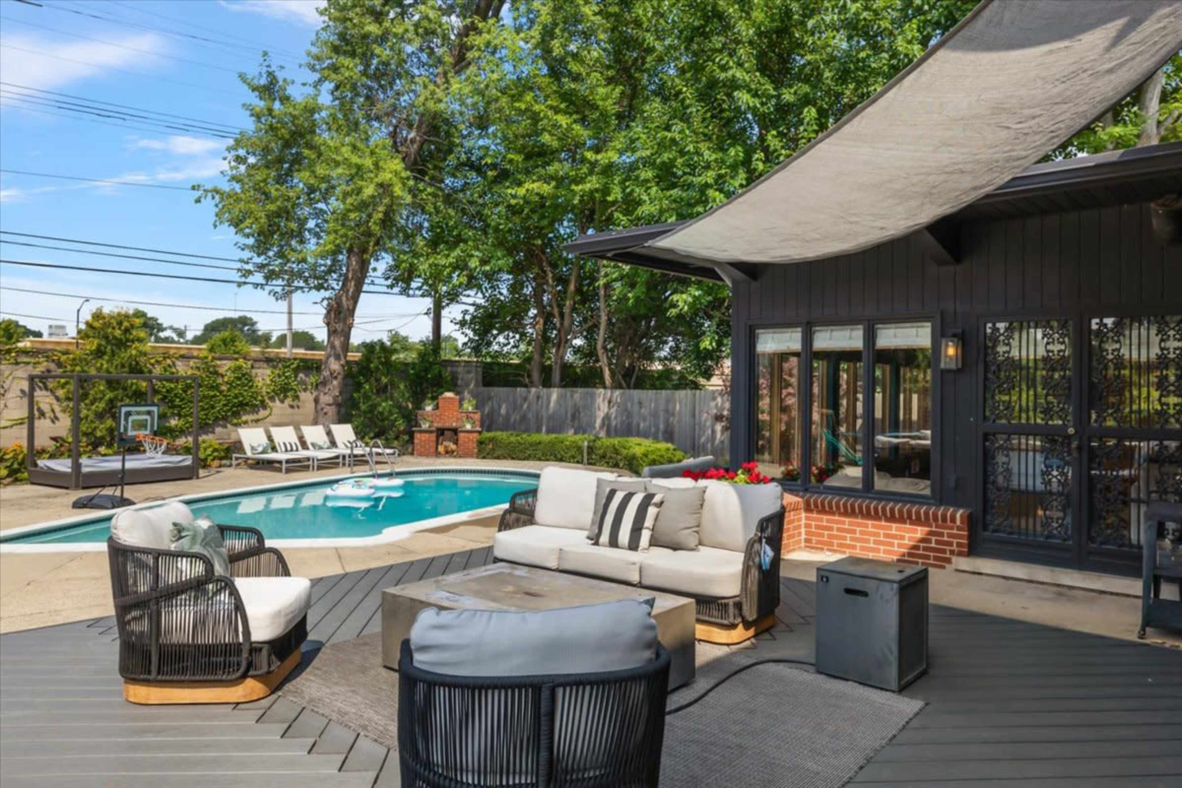 The image shows a backyard patio featuring a seating area with a circular table, overlooking a swimming pool surrounded by lounge chairs and greenery.