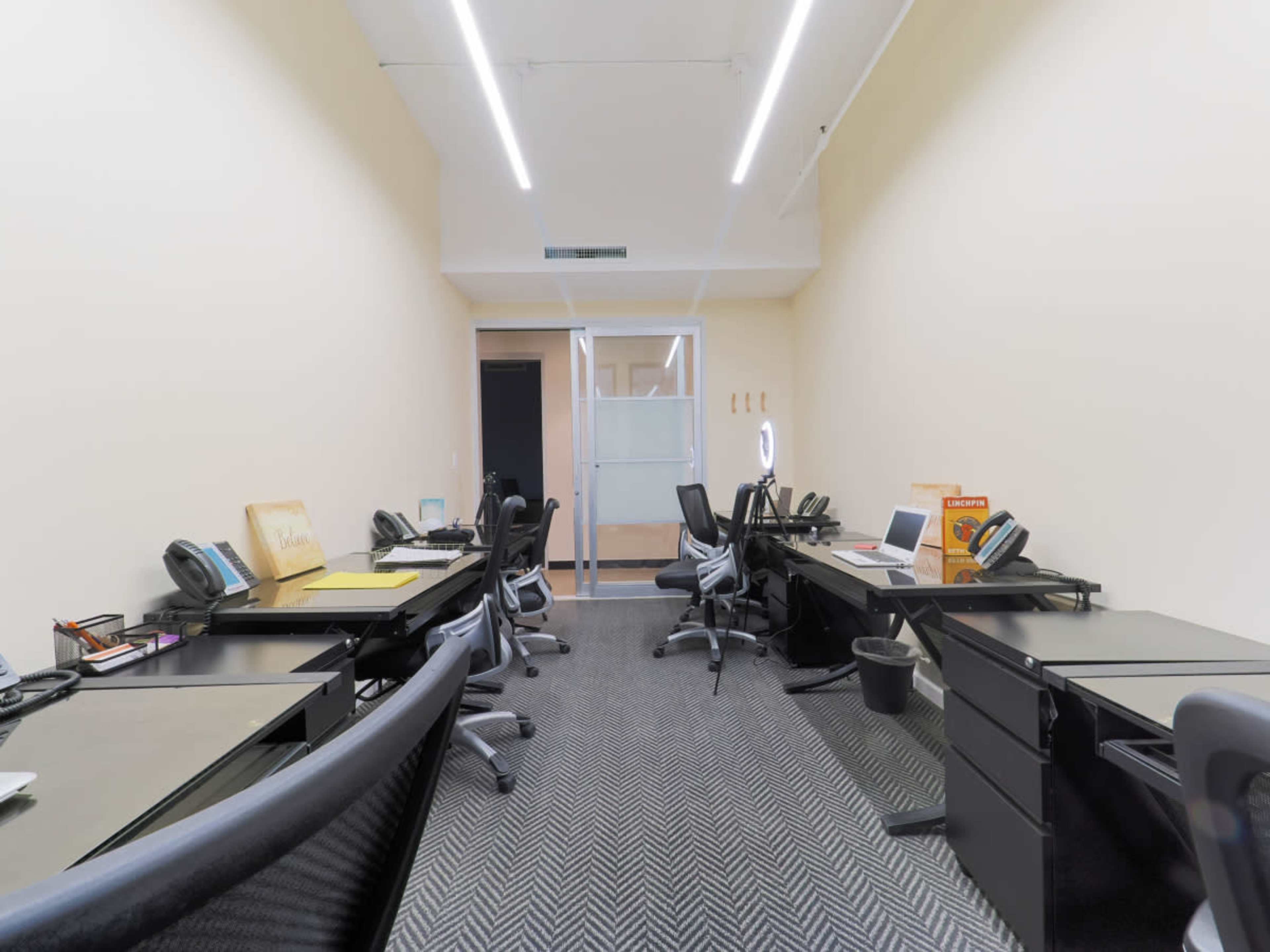 A row of black office desks with chairs, computers, and telephones arranged in a well-lit workspace.