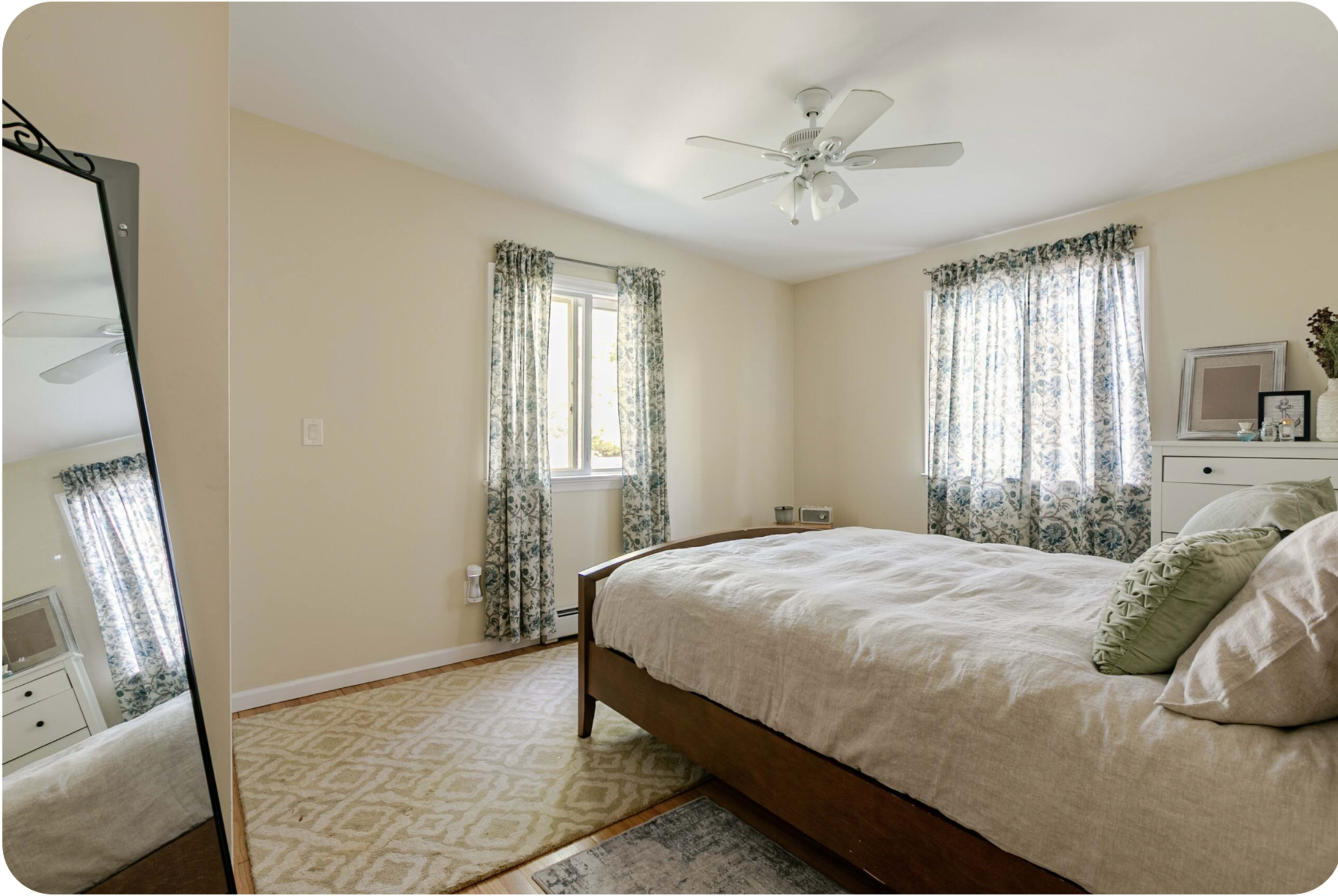 A softly lit bedroom features a bed with a light-colored comforter, a large mirror, and windows adorned with patterned curtains.