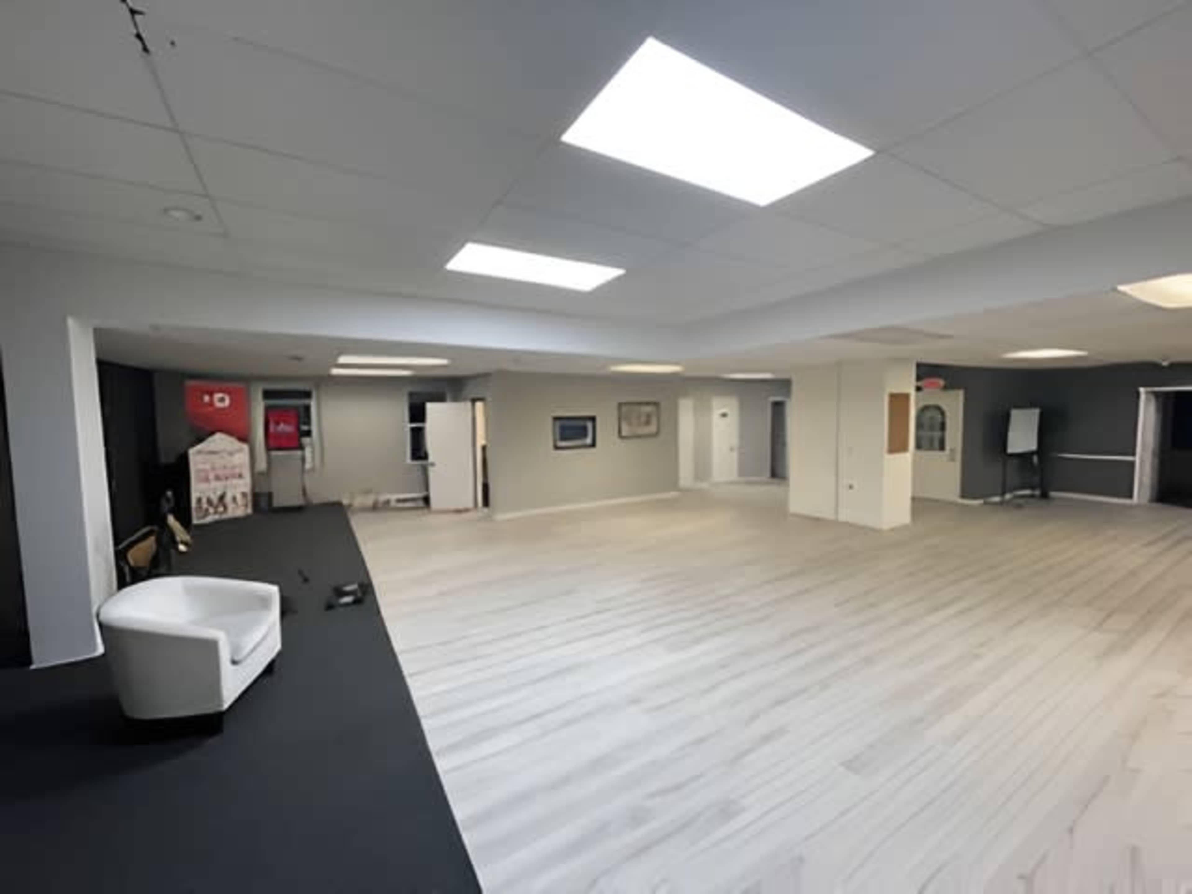 The image shows a spacious indoor area with light-colored wood flooring, white walls, and several overhead fluorescent lights.