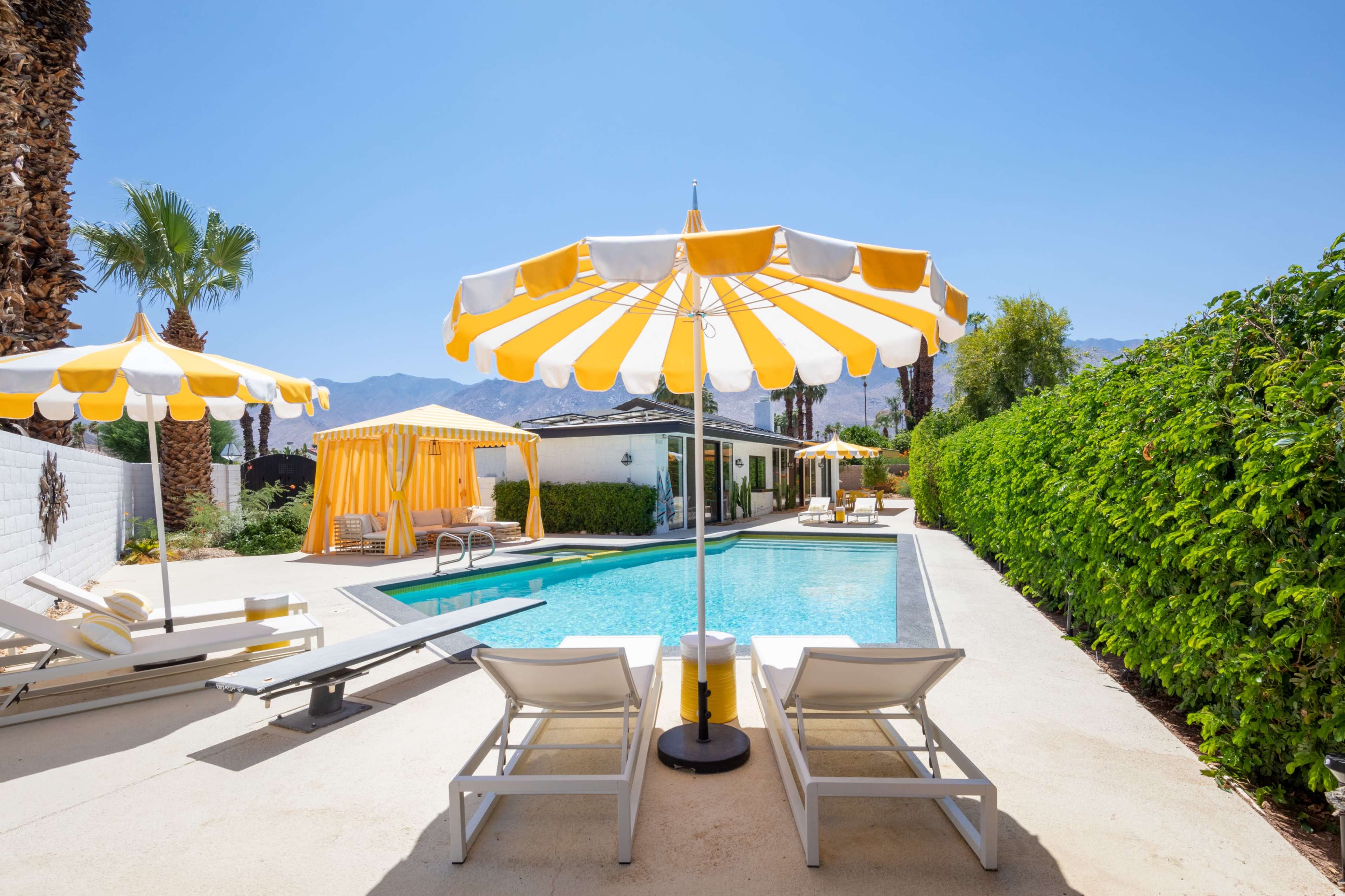 A pool area features lounge chairs and yellow and white umbrellas, surrounded by green hedges and palm trees under a clear blue sky.