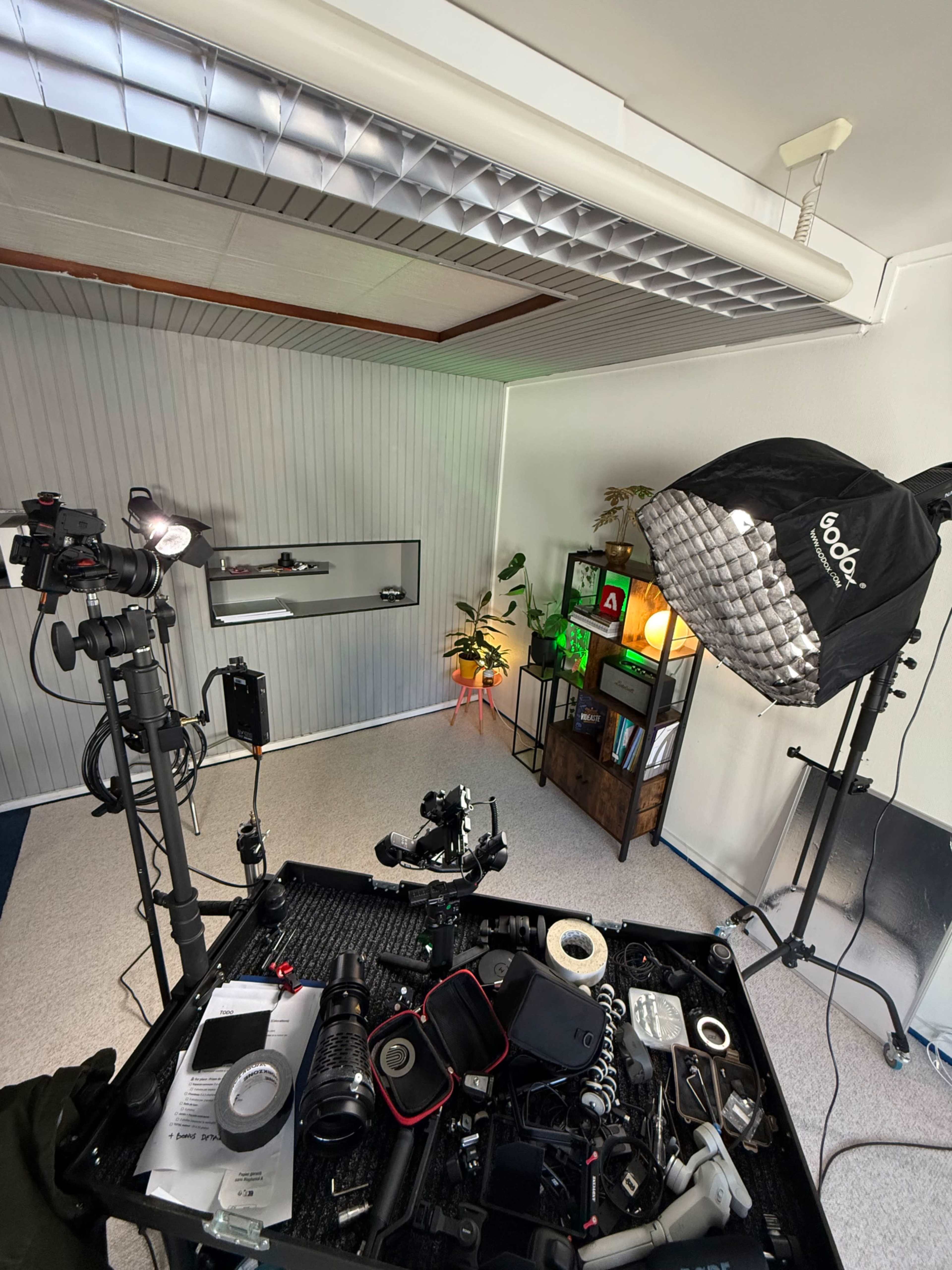 The image shows a well-equipped filming setup featuring a camera, lighting, and various filming accessories in a room with a backdrop and decorative plants.
