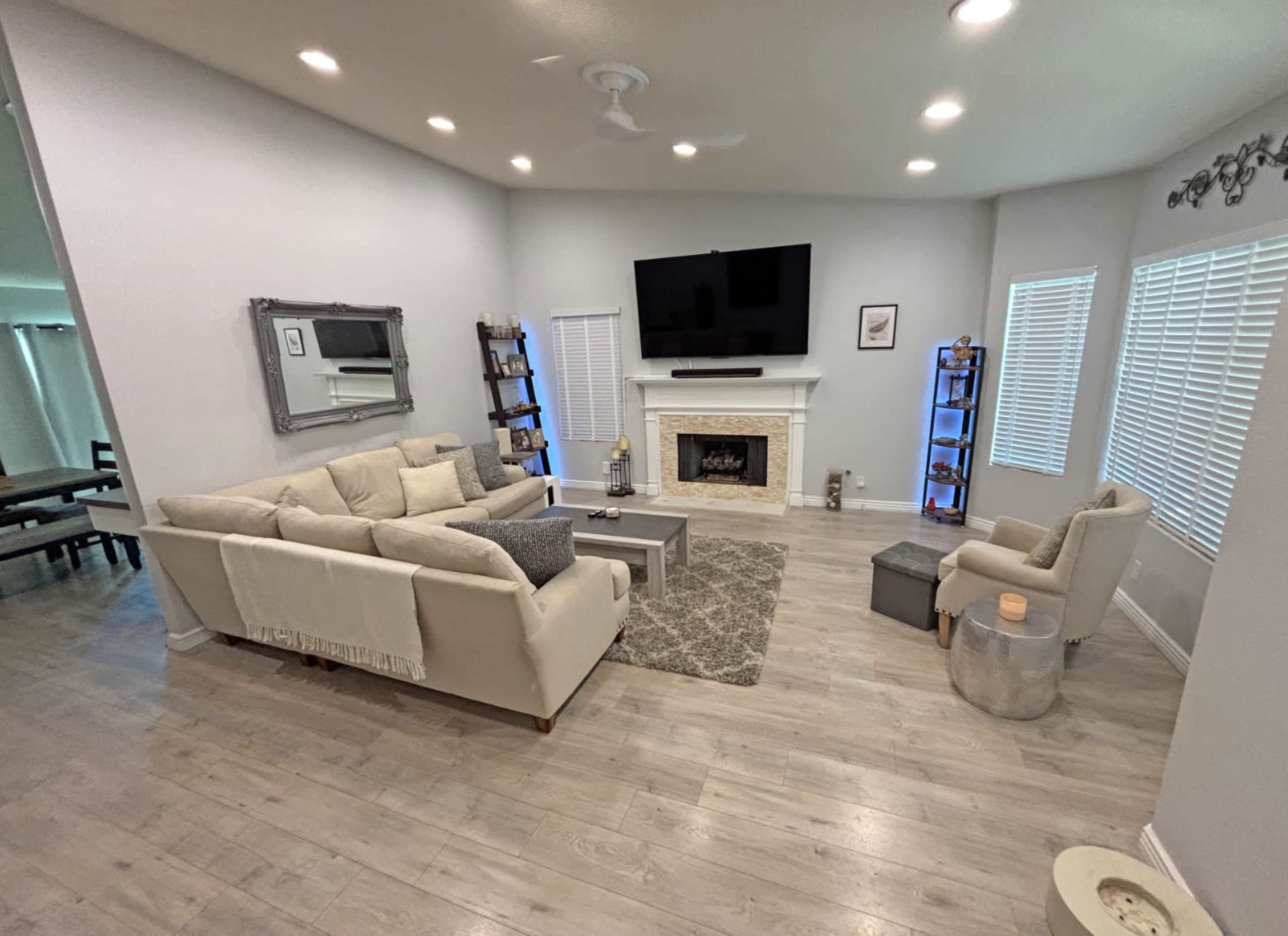 A modern living room features a light-colored sectional sofa, a coffee table, a large TV mounted above a fireplace, and bright windows with wooden blinds.
