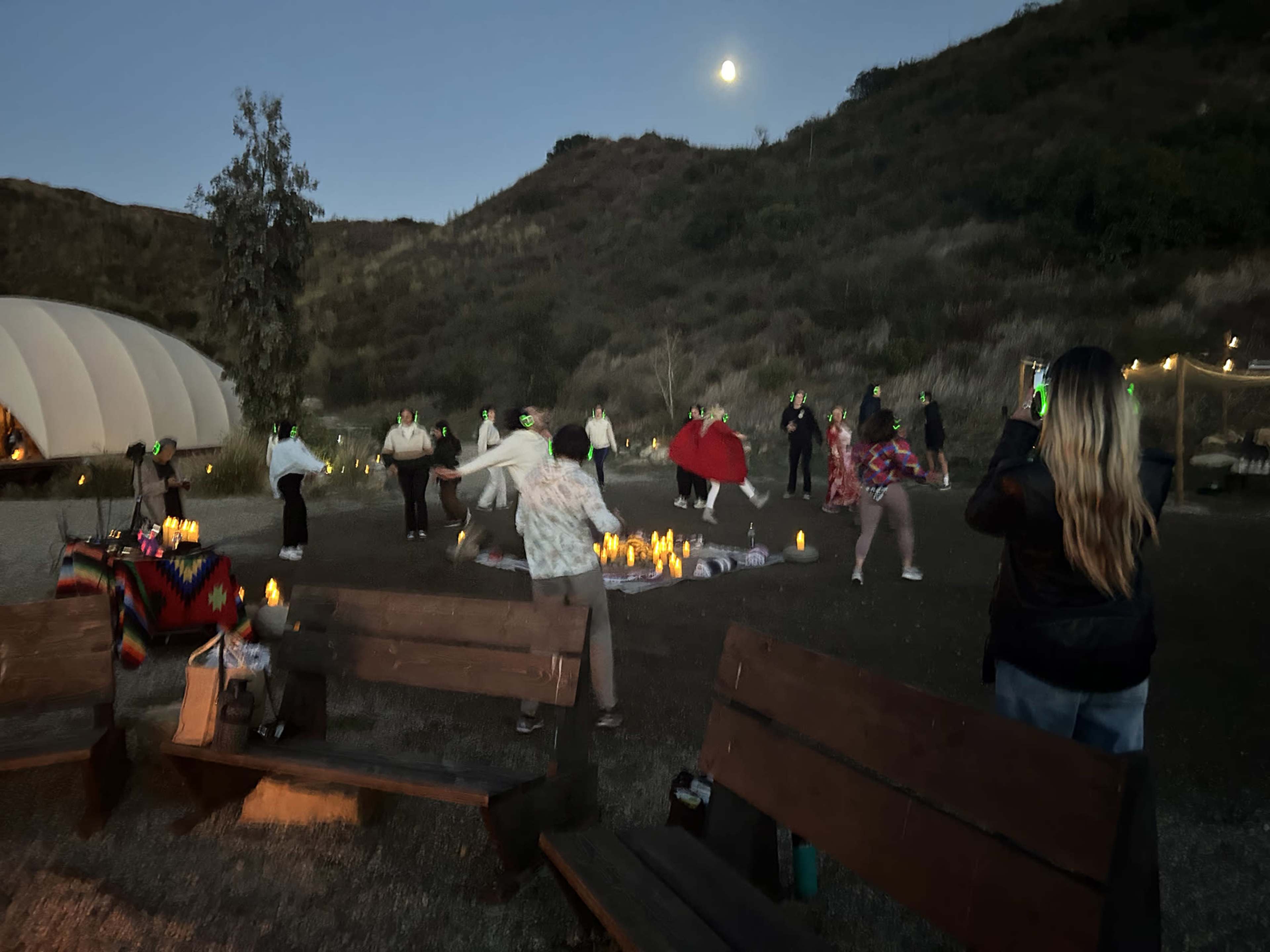 A group of people dressed in various outfits performs a dance in an outdoor setting at dusk, surrounded by candles and under a full moon.