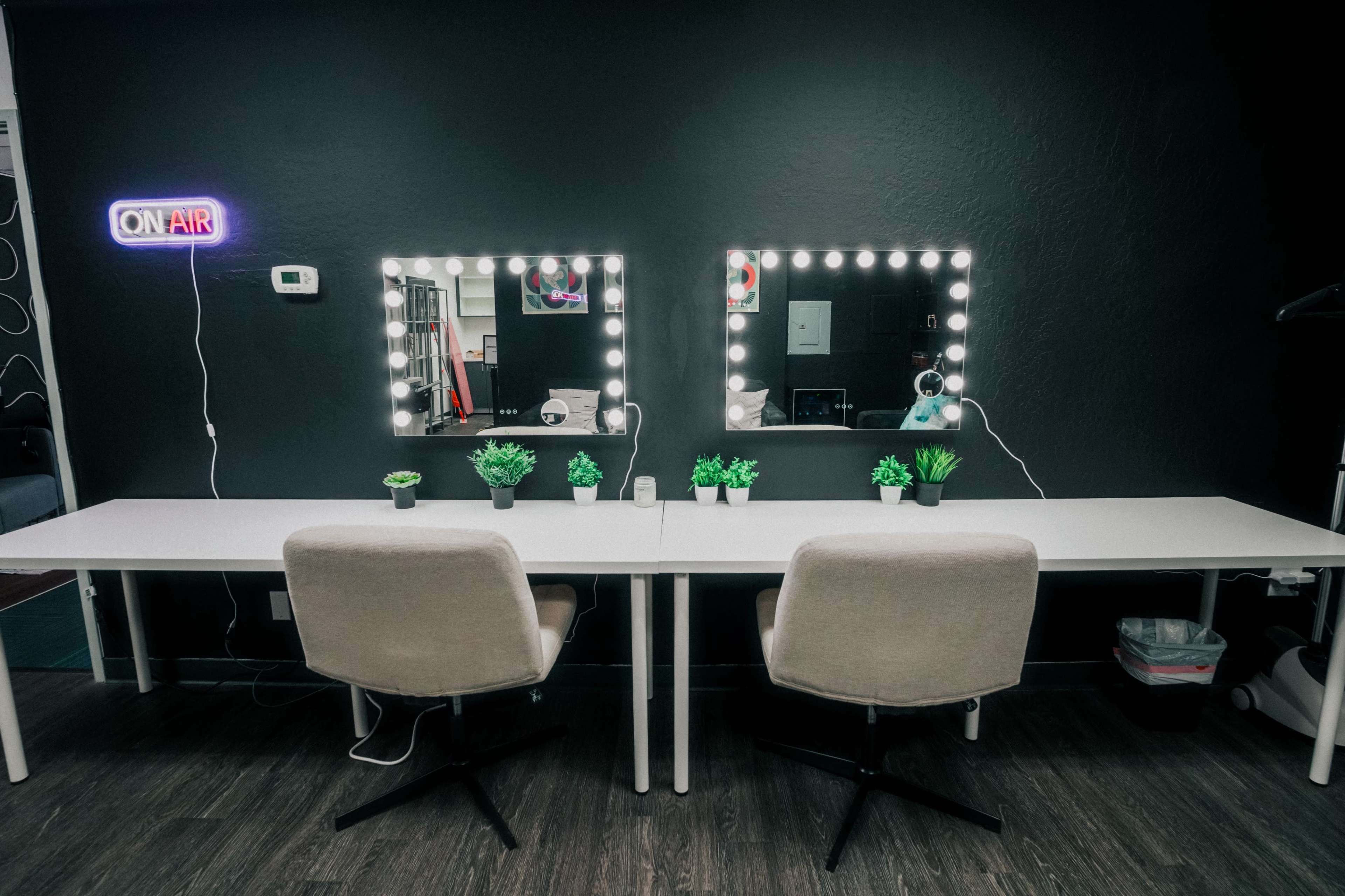 A well-lit workspace features two mirrors with lights, a long table, and small potted plants, set against a dark wall.