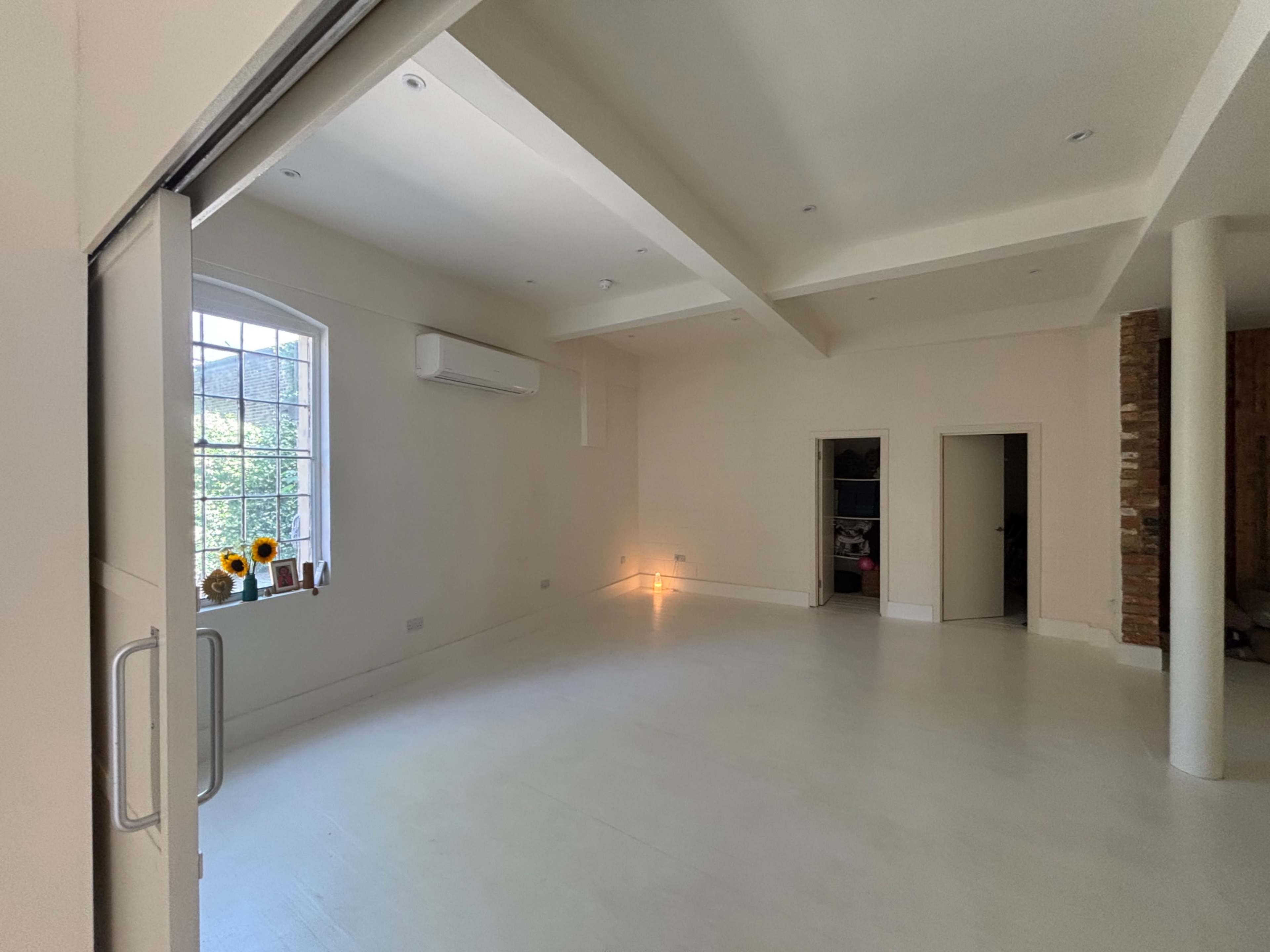 The image shows a spacious, empty room with neutral walls, a large window, and a few decorative elements such as a vase with sunflowers.