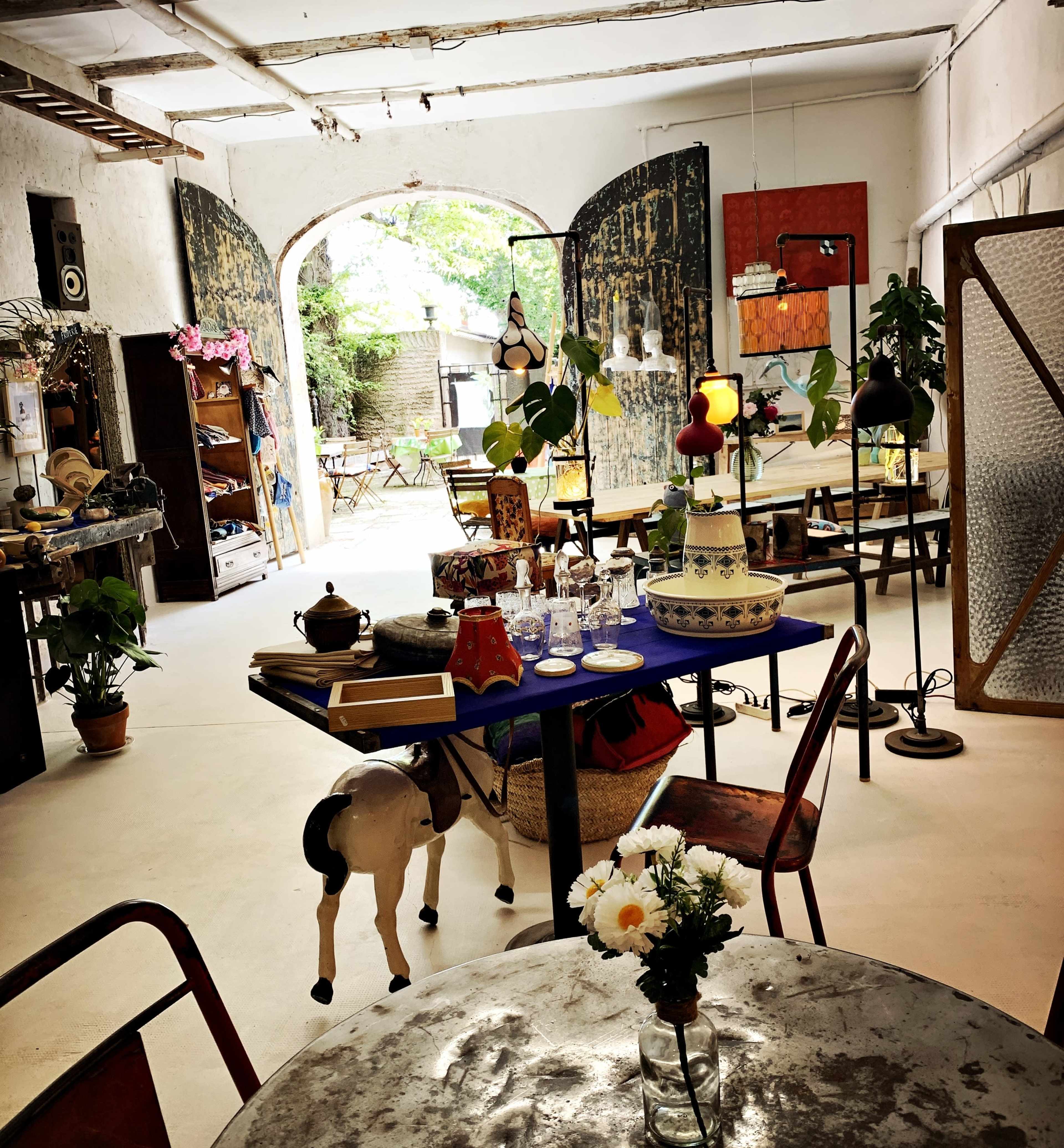 The image shows a vintage shop interior filled with various items, including furniture, lamps, and decorative objects, arranged around a central table.