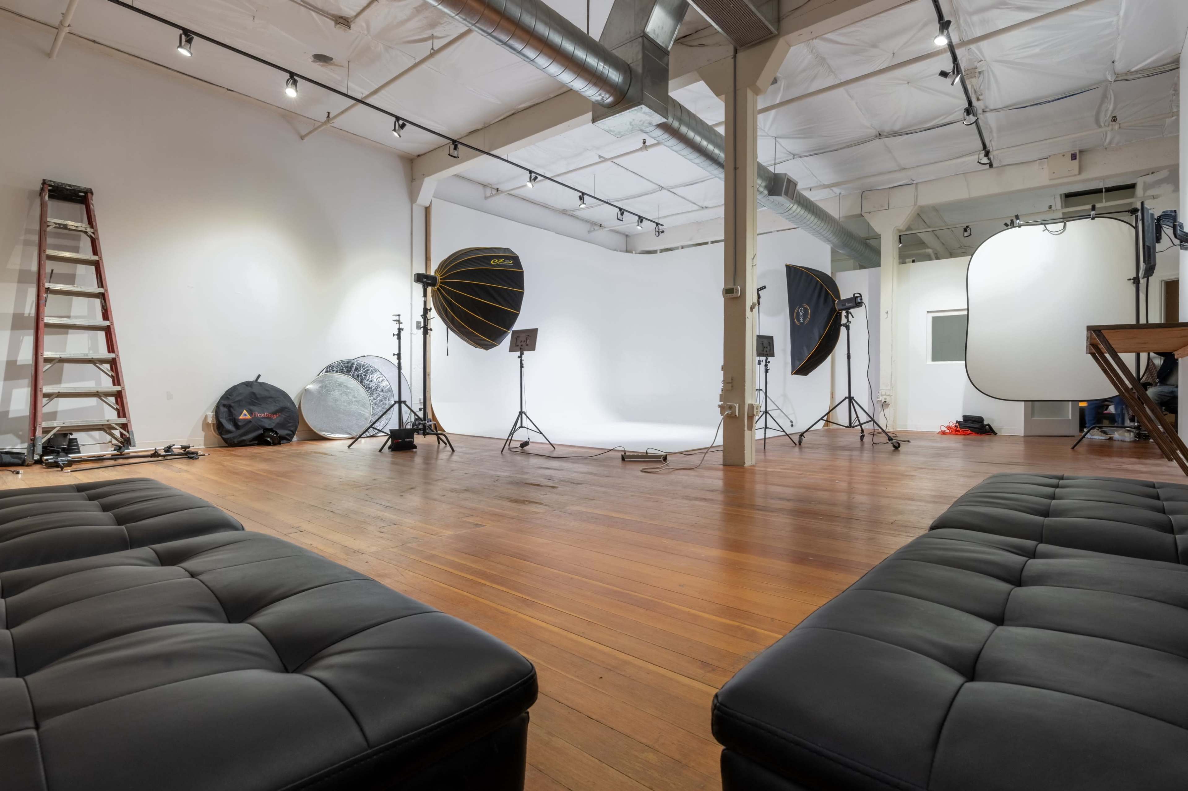 A spacious photography studio with a wooden floor, two black sofas, lighting equipment, and a white backdrop.