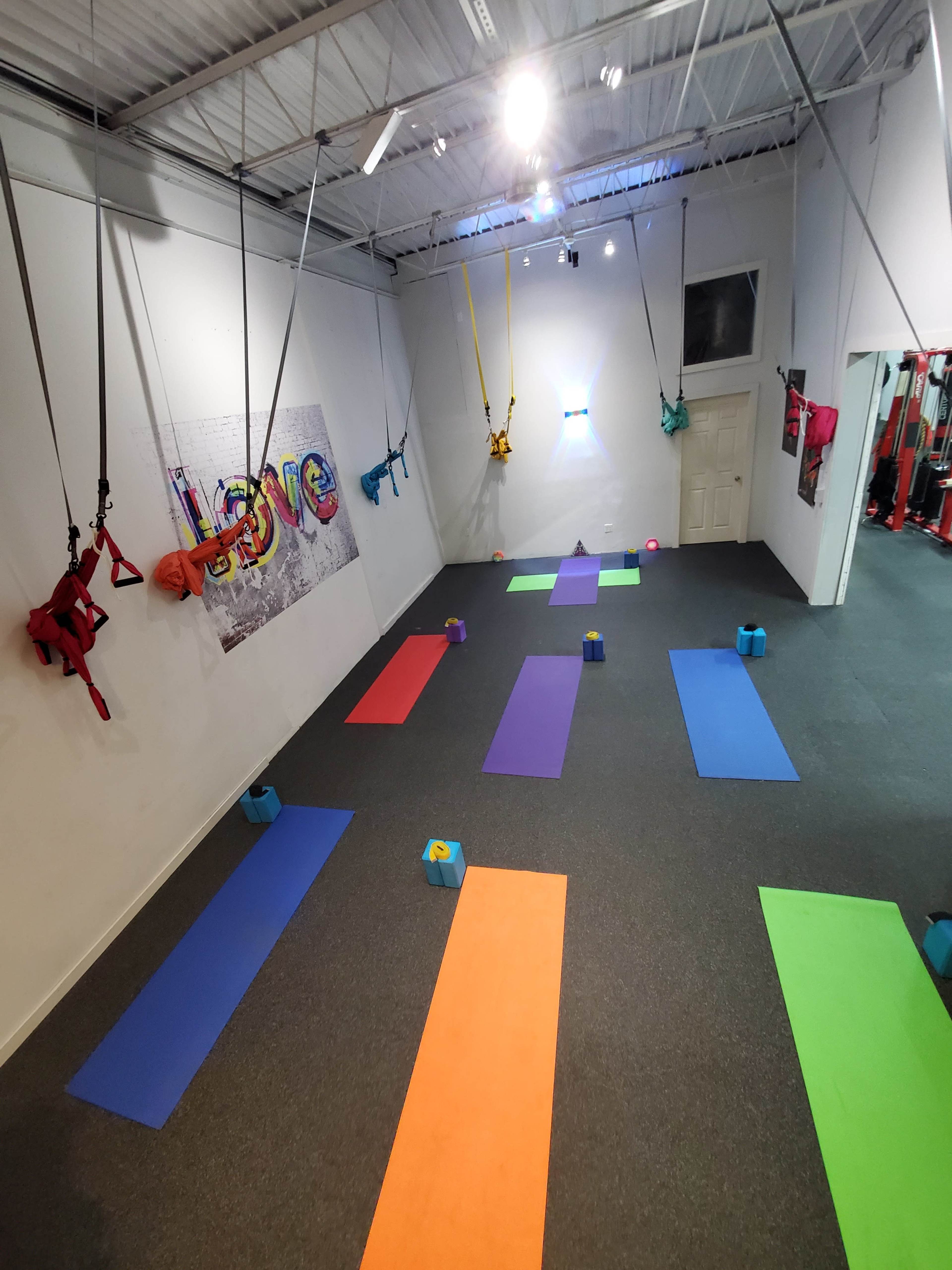A brightly colored fitness studio with various exercise mats arranged on the floor and harnesses suspended from the ceiling.