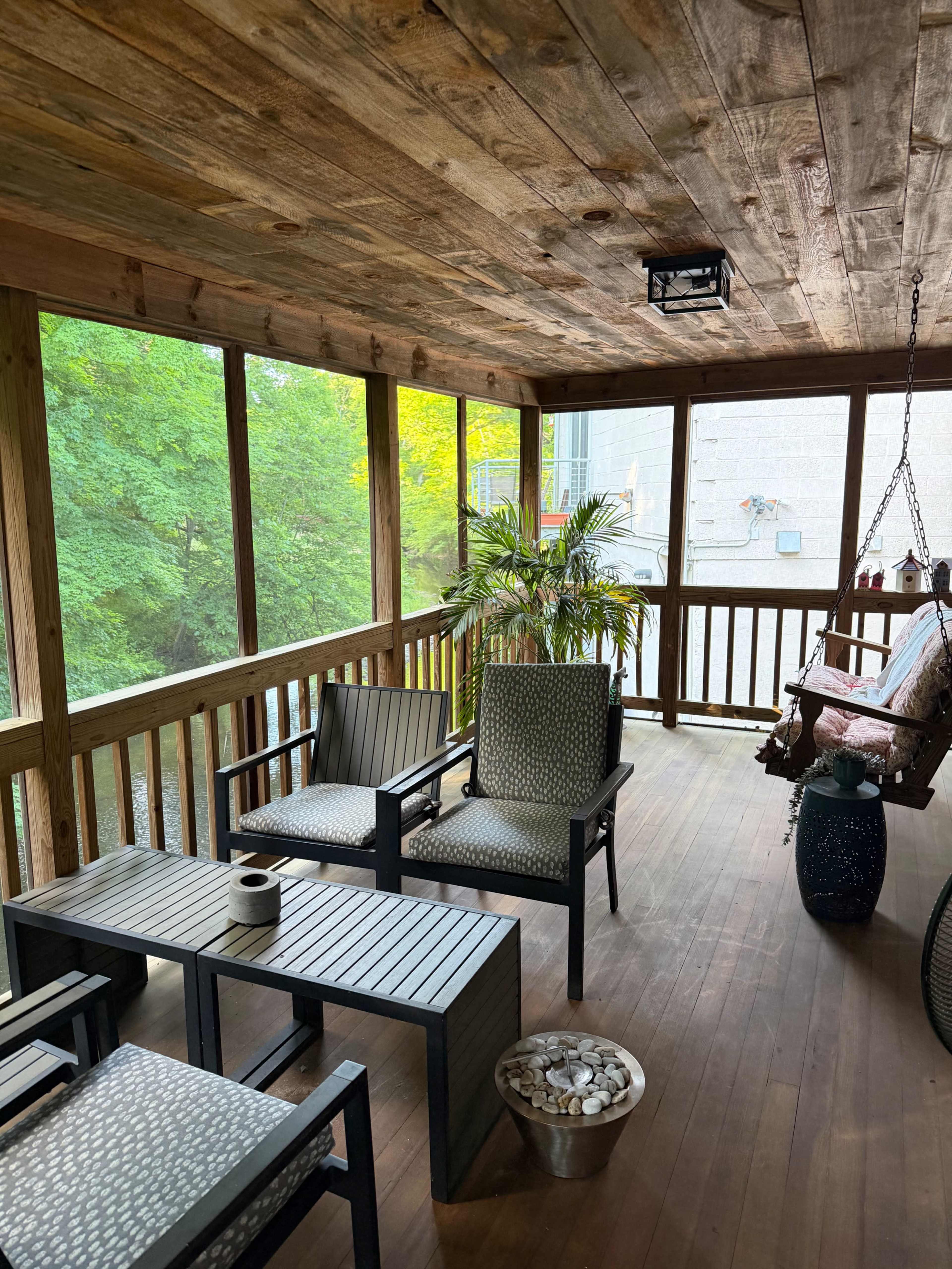 The image shows a screened-in porch with wooden walls and ceiling, featuring a seating area with black chairs, a coffee table, a potted plant, and a swing.