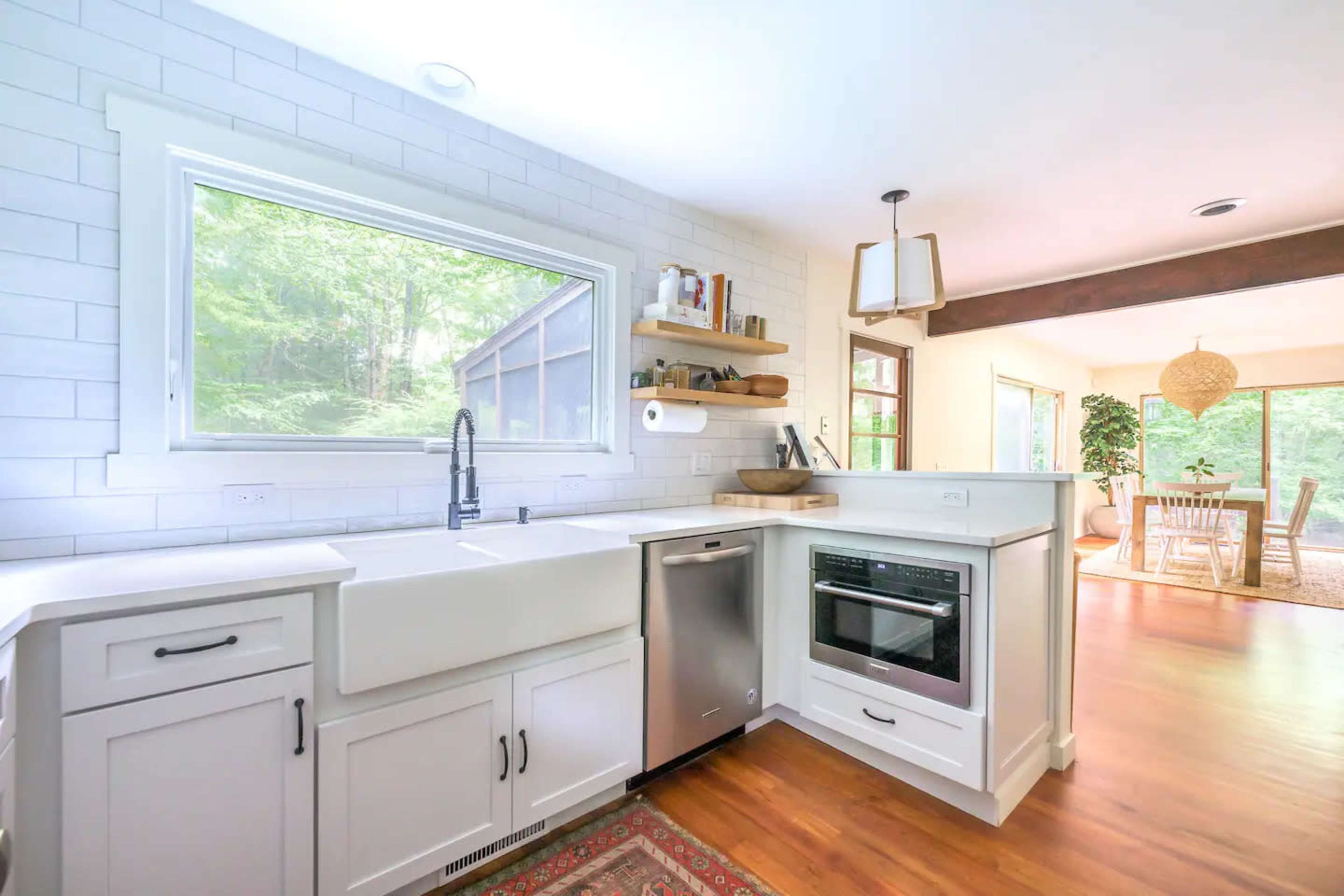 A modern kitchen with a farmhouse sink, stainless steel appliances, white cabinetry, and a window overlooking greenery.