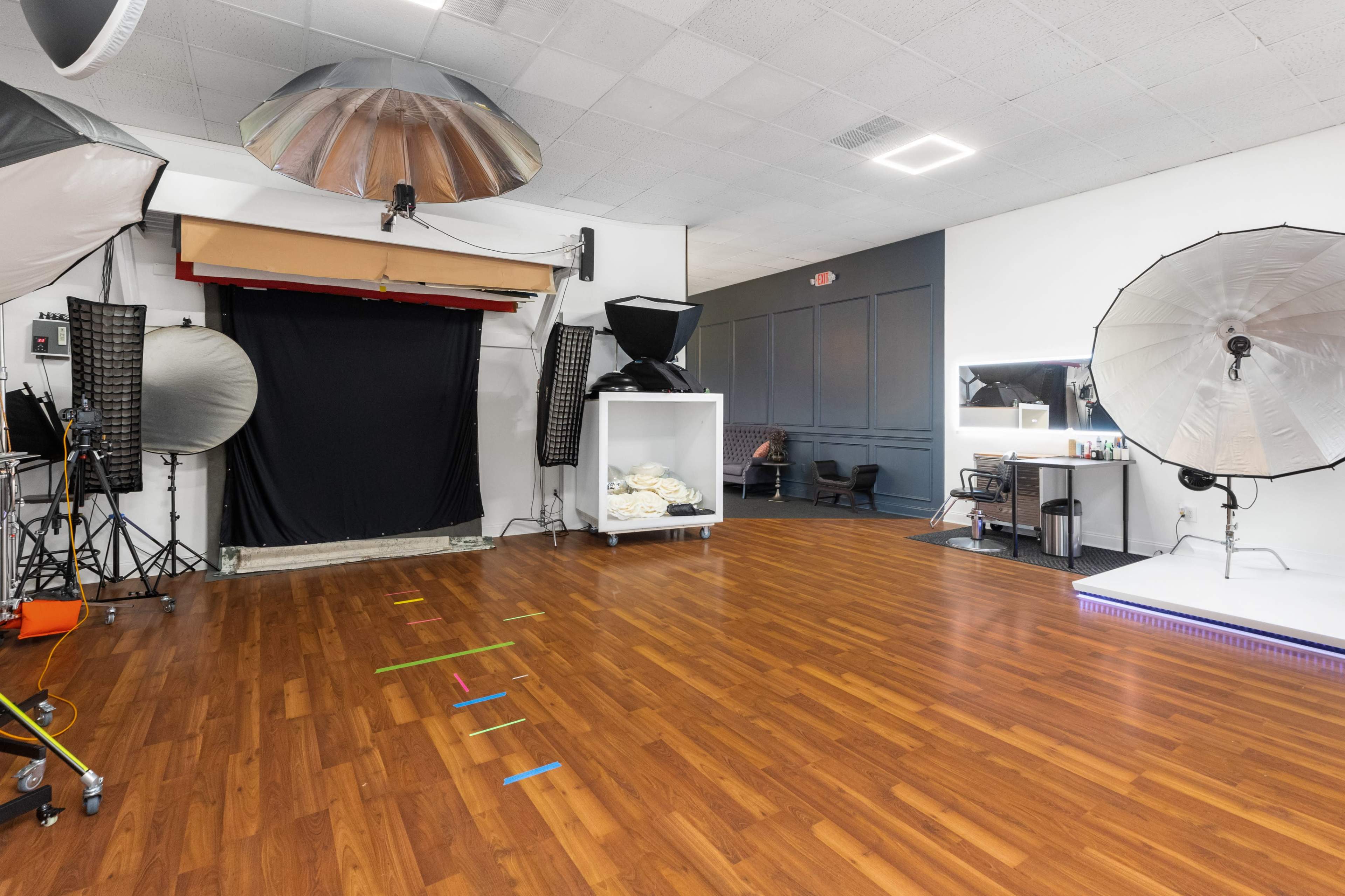 The image shows a photography studio with a wooden floor, various lighting equipment, a backdrop area, and a seating area.