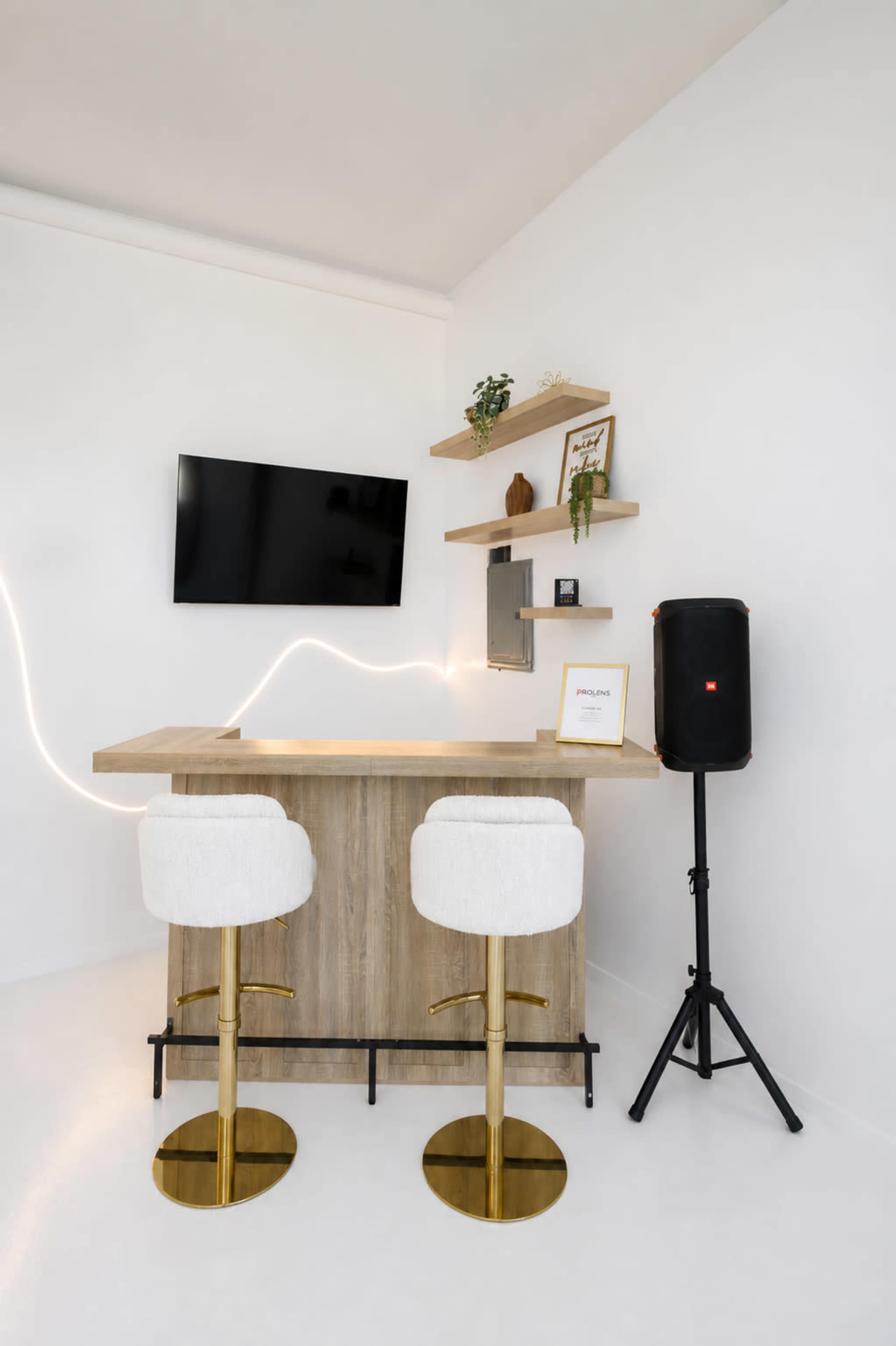 The image features a modern home bar with two cushioned stools, a wooden counter, a wall-mounted TV, and a speaker on a stand.